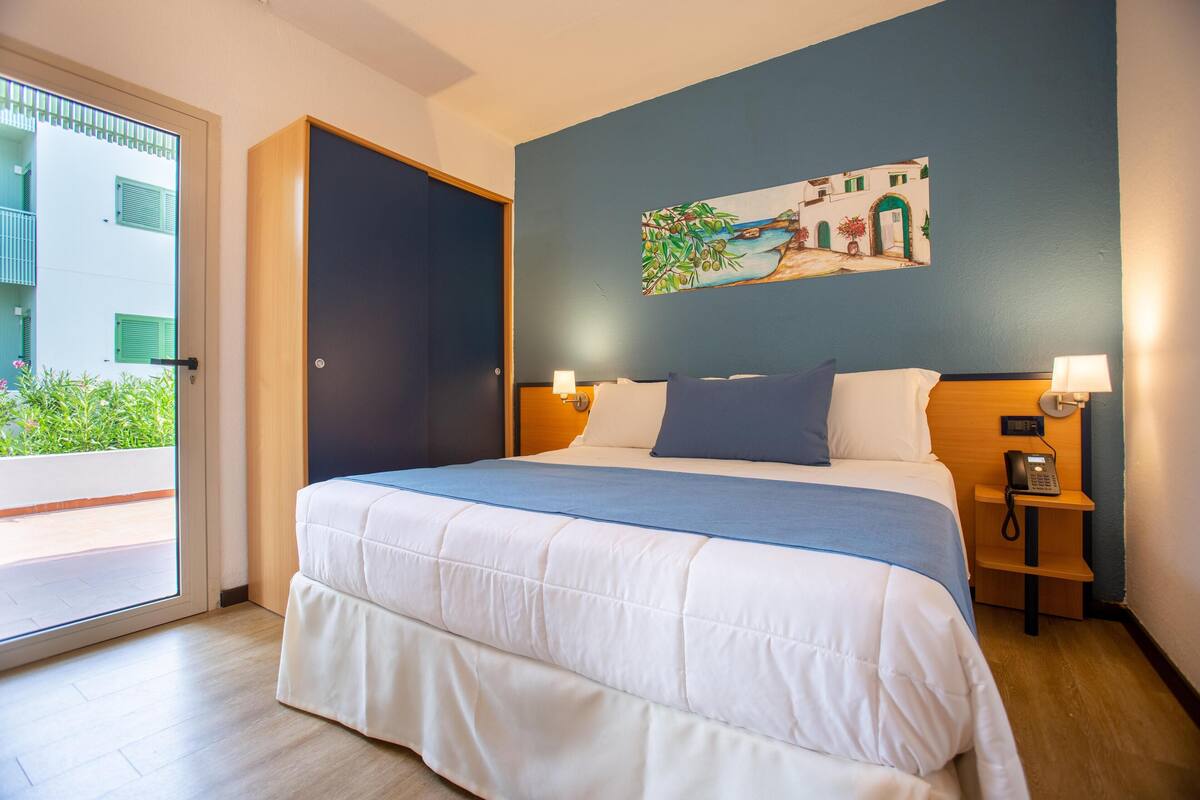 A hotel room with a bed, a painting on the wall, a door, and a nightstand with a phone.