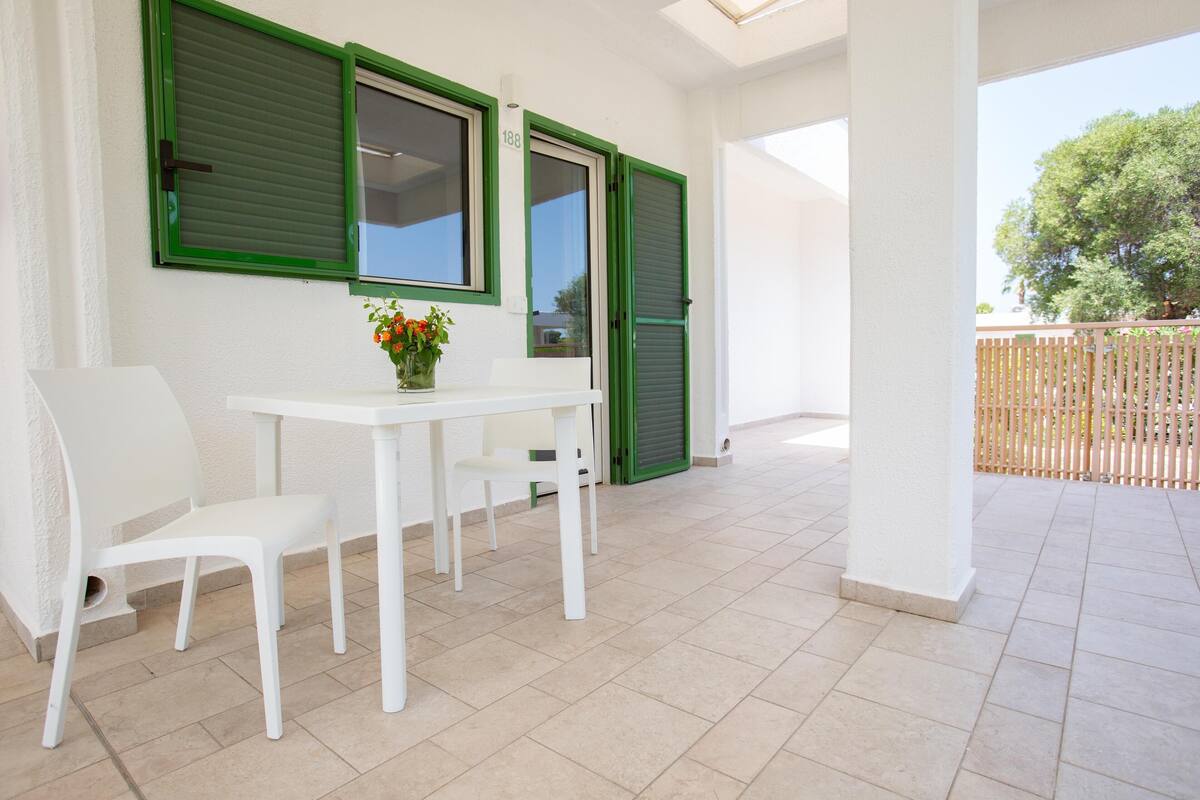 A clean, white interior with a table and chairs, a small vase with flowers, and green shutters on the windows.