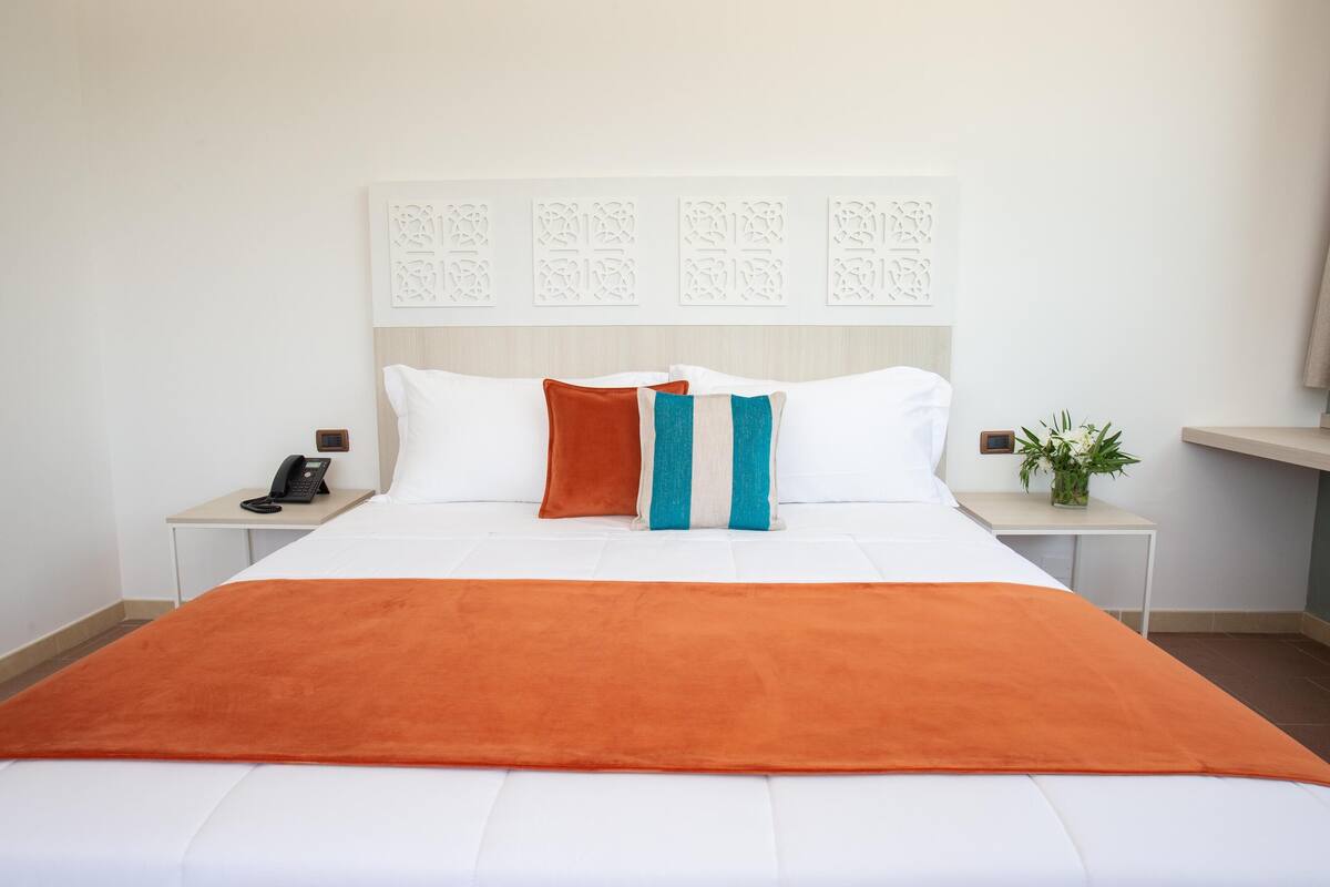 A neatly made bed with white and orange bedding, two pillows, and a bedside table with a phone and a small plant.