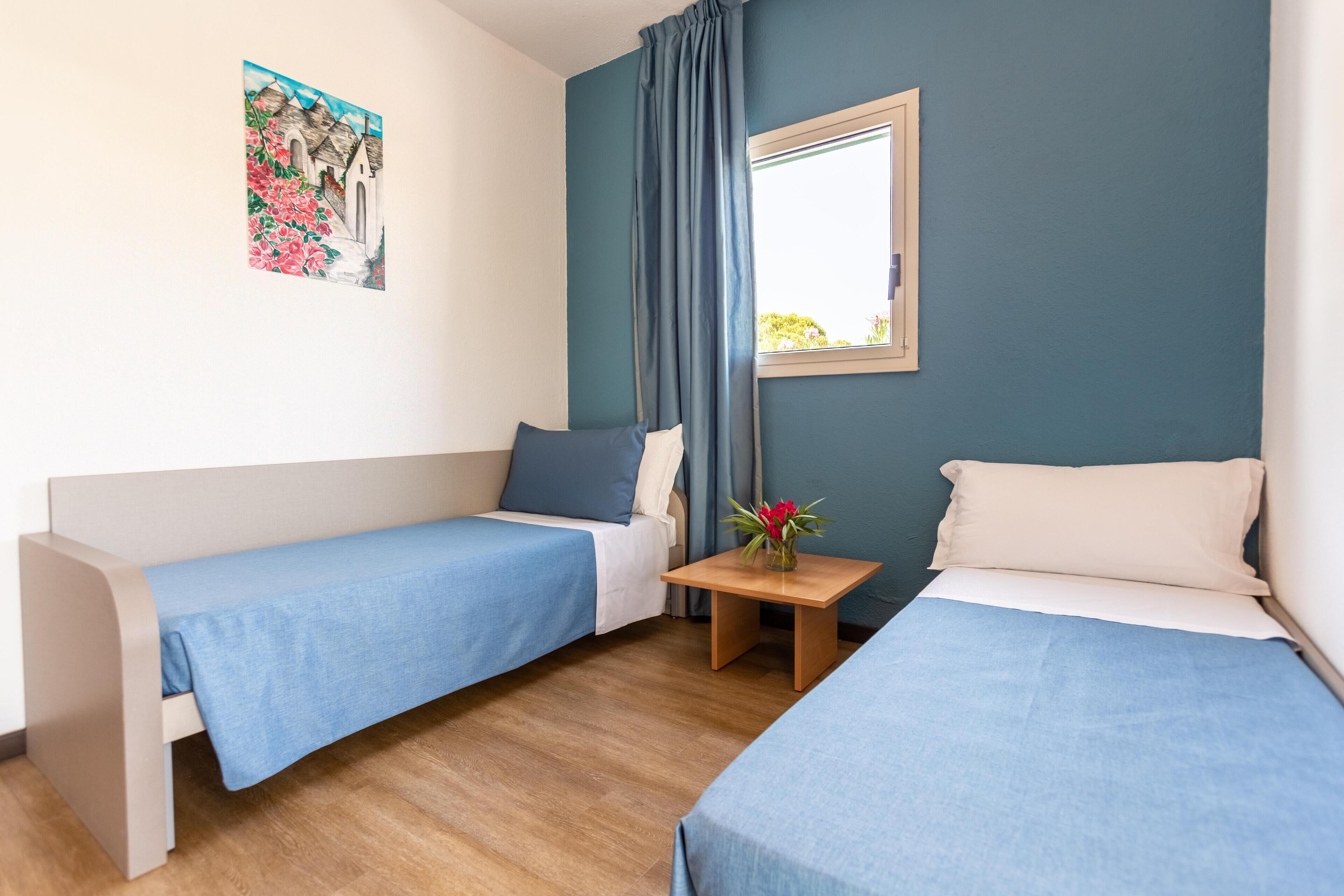 A room with two single beds, a small table with a vase of flowers, a window with curtains, and a painting on the wall.