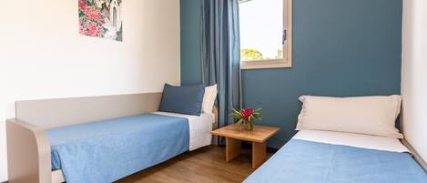 A room with two single beds, a small table with a vase of flowers, a window with curtains, and a painting on the wall.