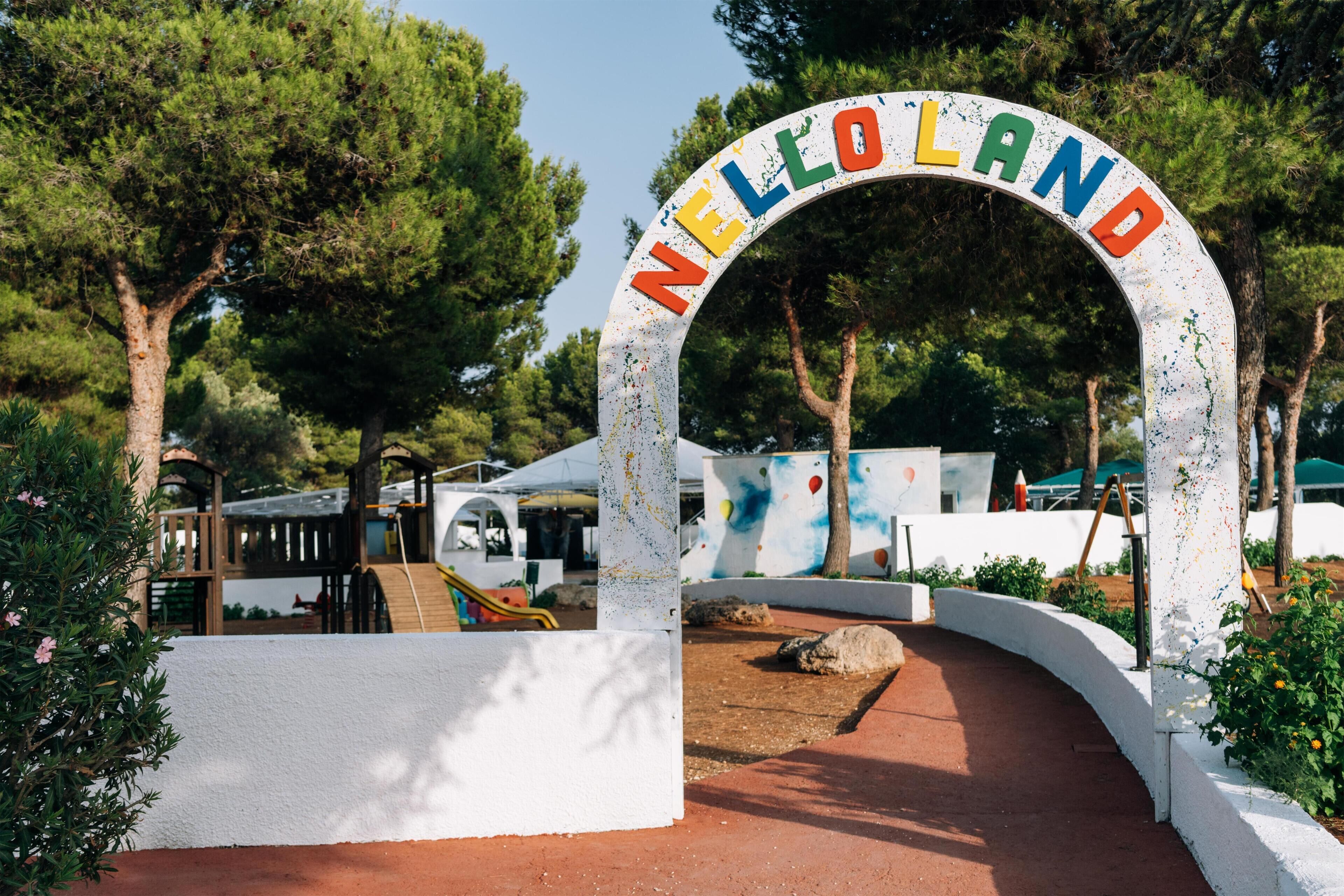 Un arco bianco con la scritta "NELLOLAND" in lettere colorate, che dà accesso a un vialetto alberato con un'area giochi sullo sfondo.