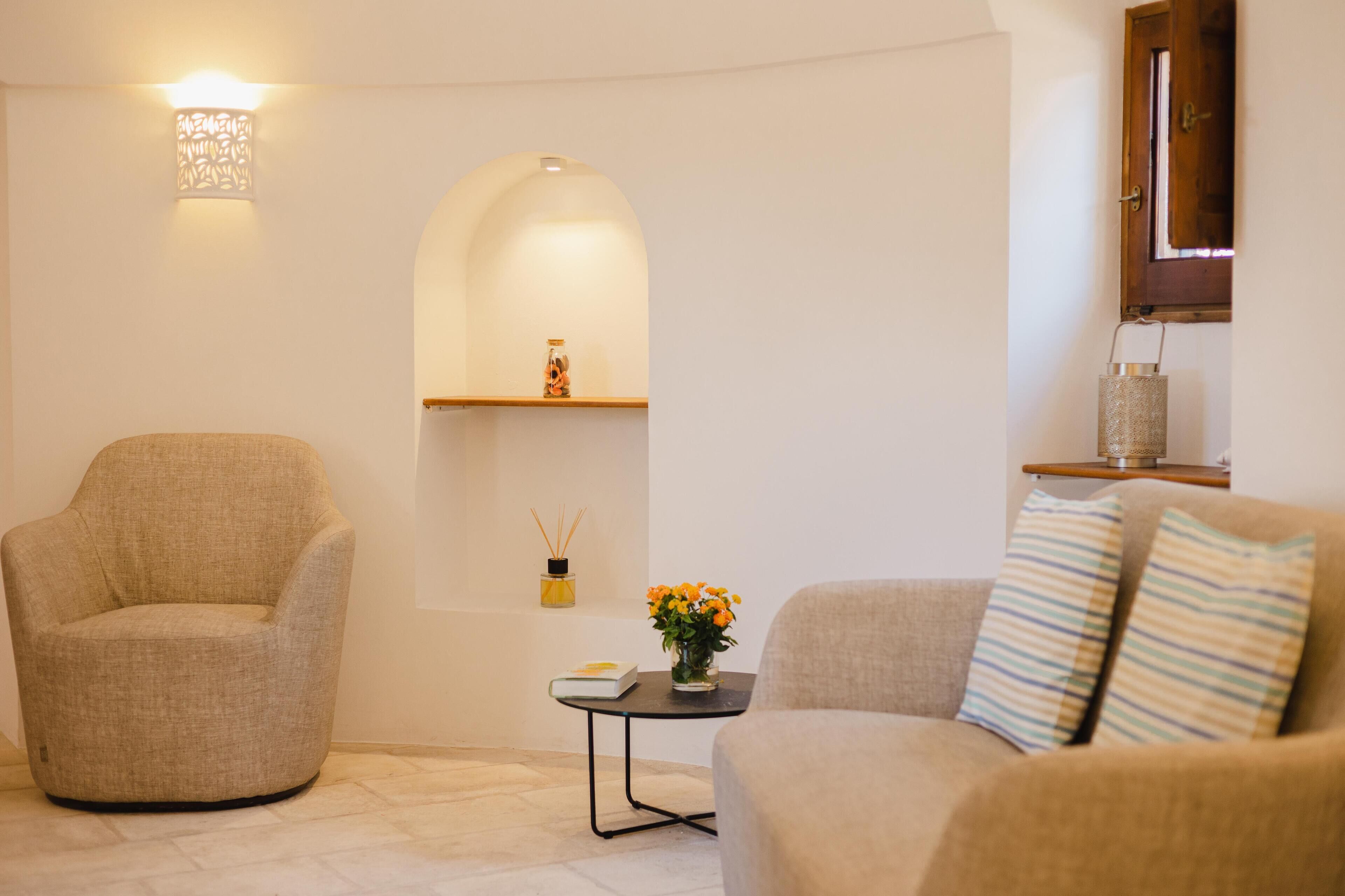 Un salon avec un fauteuil beige, un canapé agrémenté de coussins à rayures, une petite table sur laquelle est posé un vase de fleurs, et une étagère murale ornée d’objets décoratifs.
