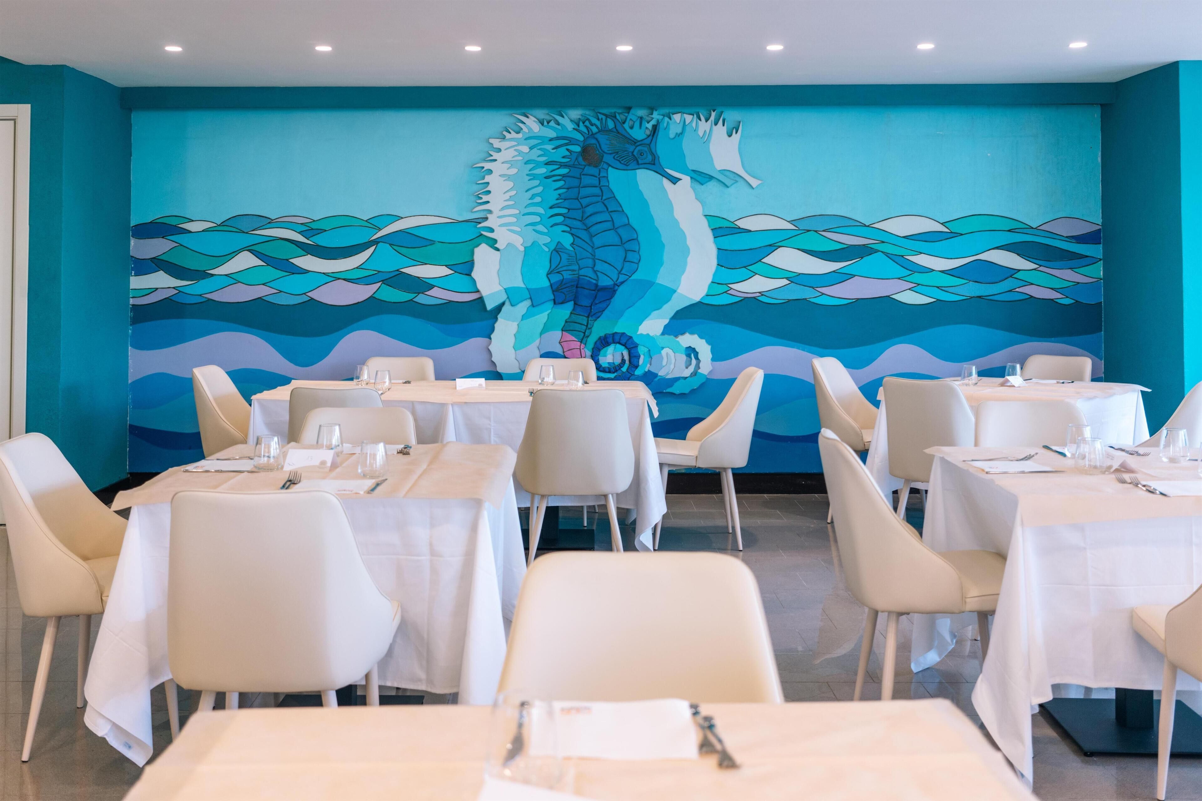 Un comedor con mesas dispuestas para el servicio, un mural de caballito de mar en la pared, y una pared color aguamarina.