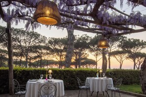 Un espace de restauration en plein air, avec des tables disposées sous une pergola ornée de glycines violettes, le tout entouré d’arbres et d’une haie.