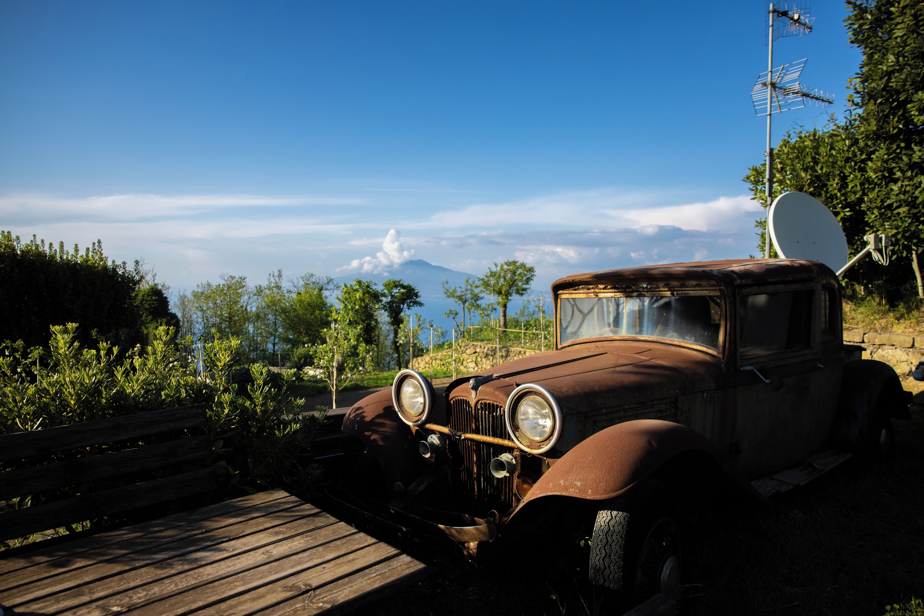 An old car parked near a wooden bench with a mountain in the background.