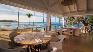 Un restaurant situé en bord de mer, avec des tables et des chaises en bois, de grandes fenêtres donnant sur l’océan et un ventilateur de plafond.