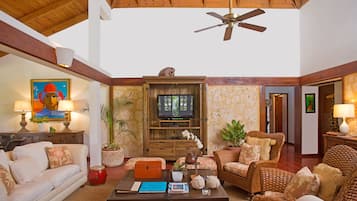 A spacious living area with a ceiling fan, a television, wicker furniture, and a coffee table with books and decorative items.