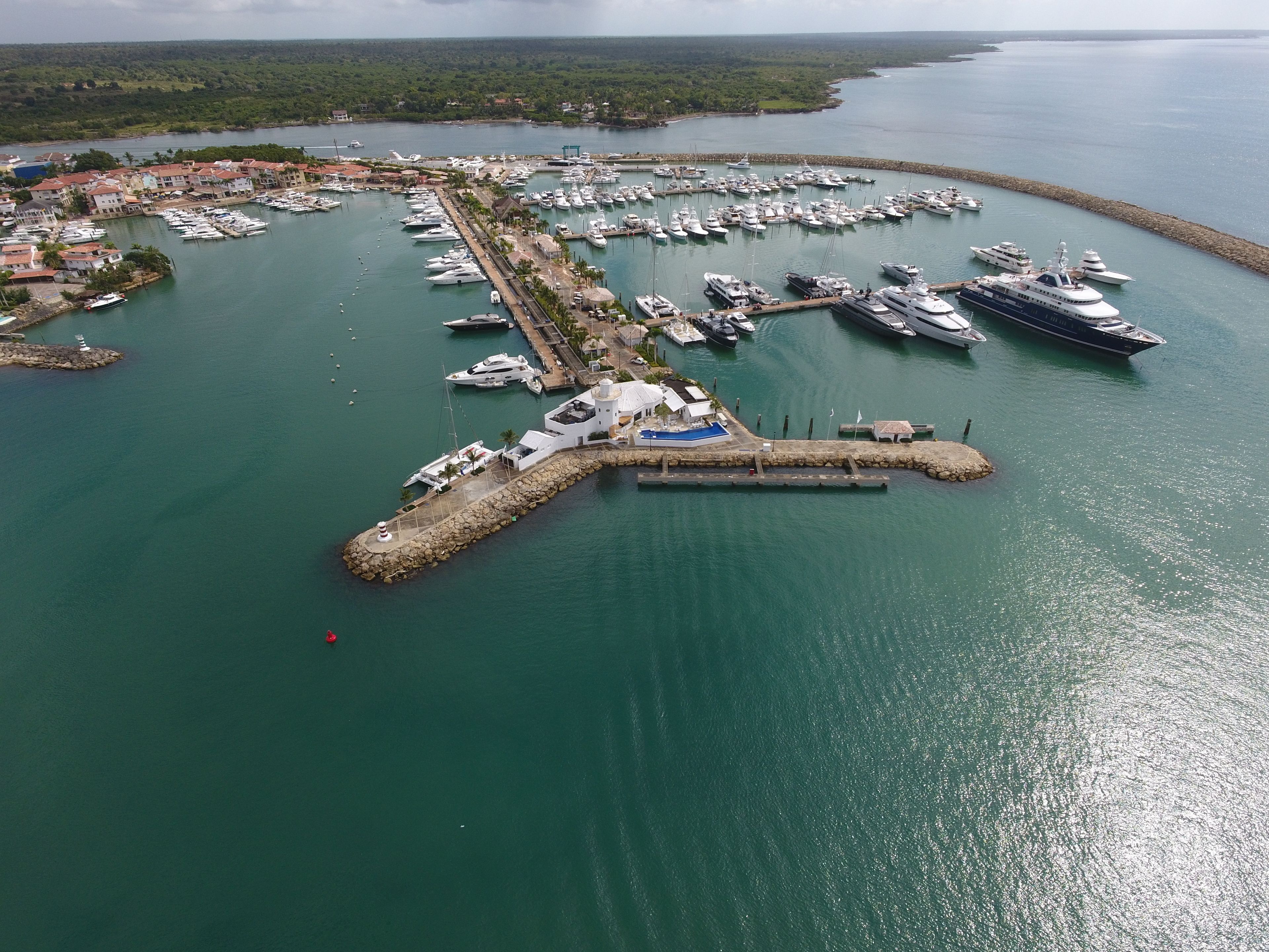 Vista aérea de un puerto deportivo con numerosas embarcaciones amarradas, una pequeña isla con un faro y una costa boscosa al fondo.