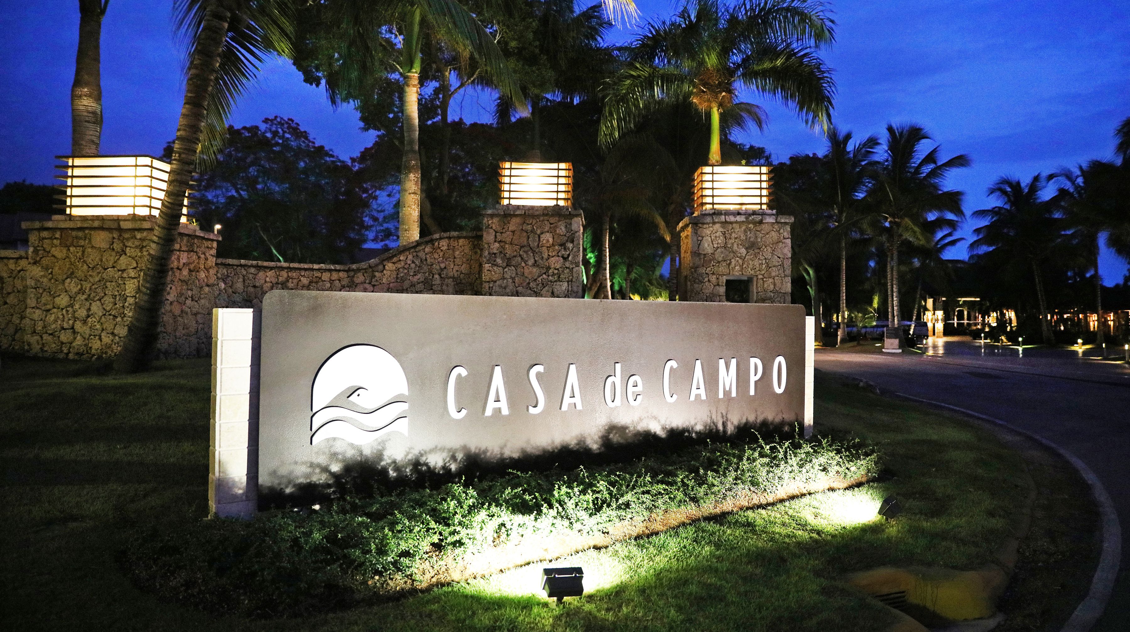 Senyal per a “CASA de CAMPO” amb un logotip, davant d'un fons de palmeres i murs de pedra, il·luminat per una llum ambiental.