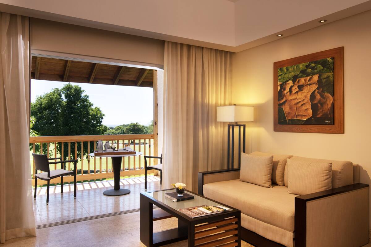 Habitación de hotel con sofá, una mesita, una lámpara y un balcón con vistas a árboles.