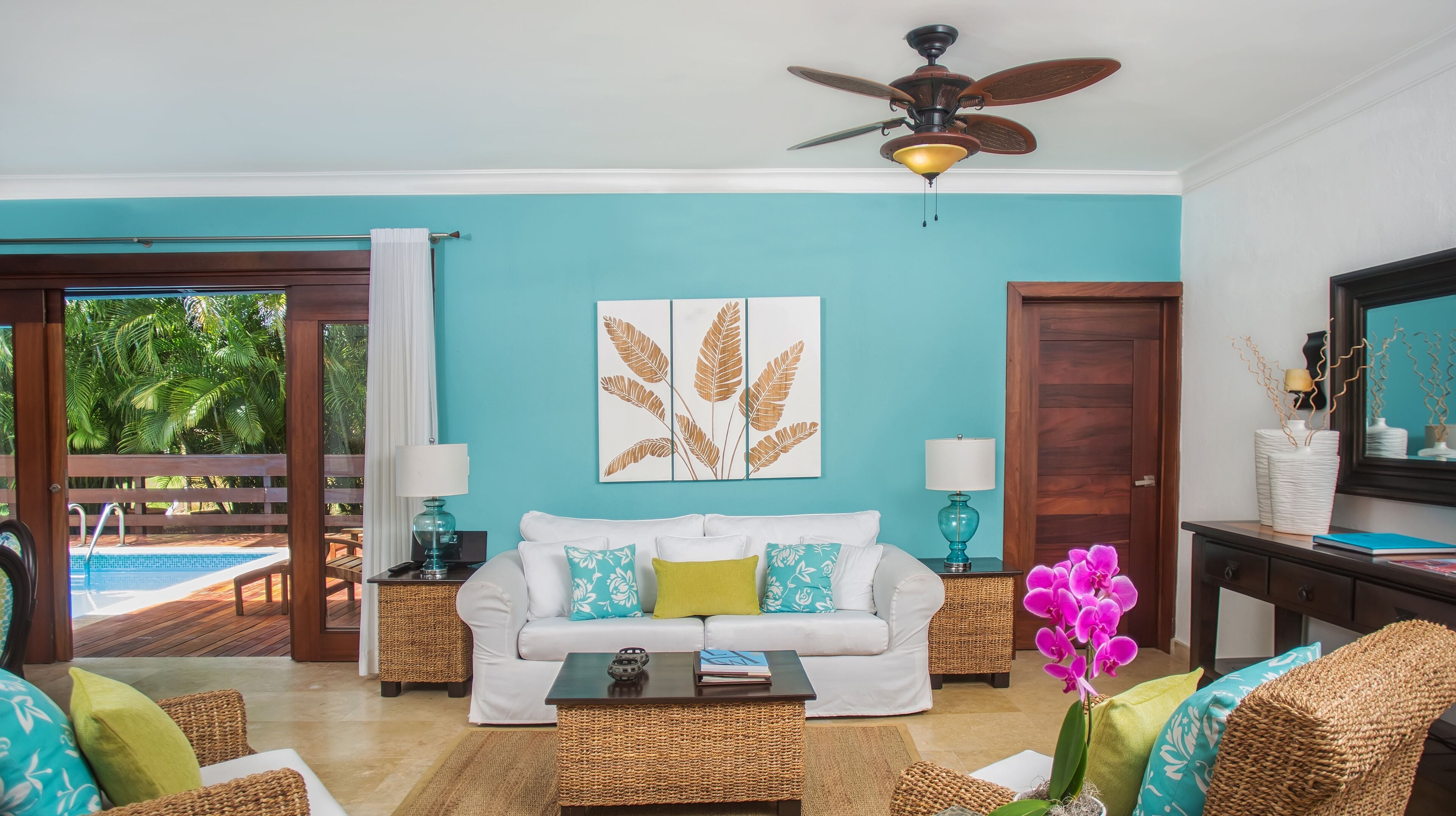 A living room with a white sofa, teal cushions, a ceiling fan, and a large window overlooking a pool.