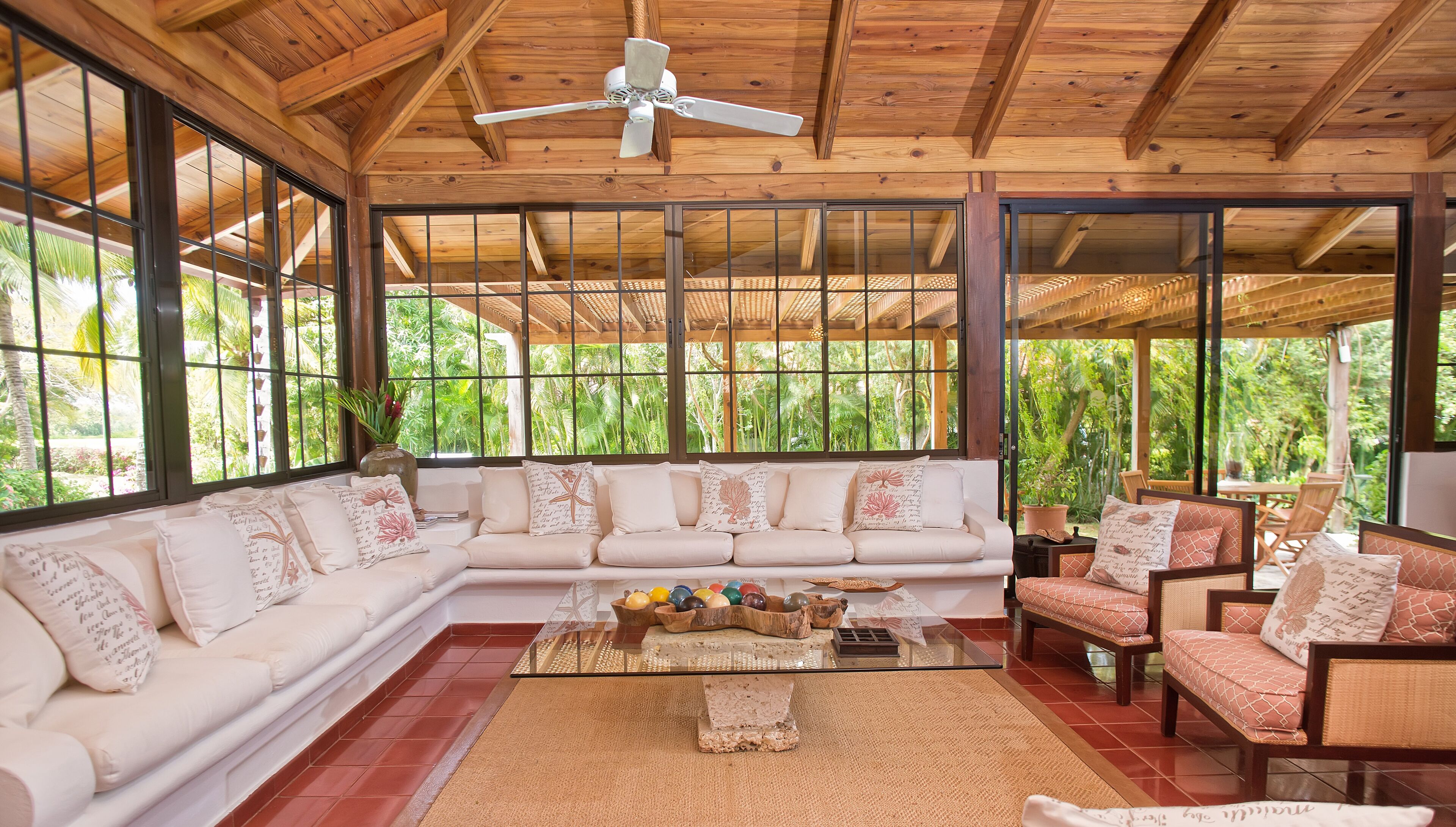 A spacious room with a large wooden ceiling, a white sofa with patterned cushions, a glass coffee table, and a view of lush greenery outside.