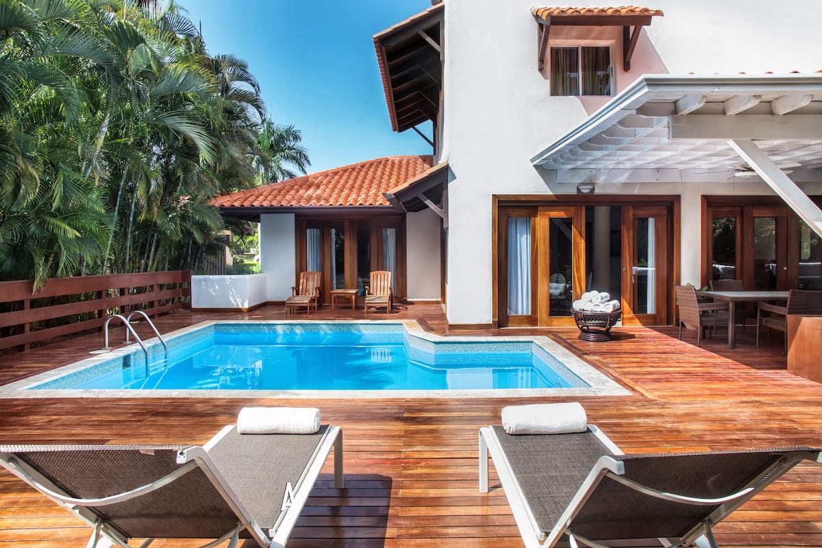 Zona de piscina con sillones de descanso, una terraza de madera y un edificio con tejado de tejas.