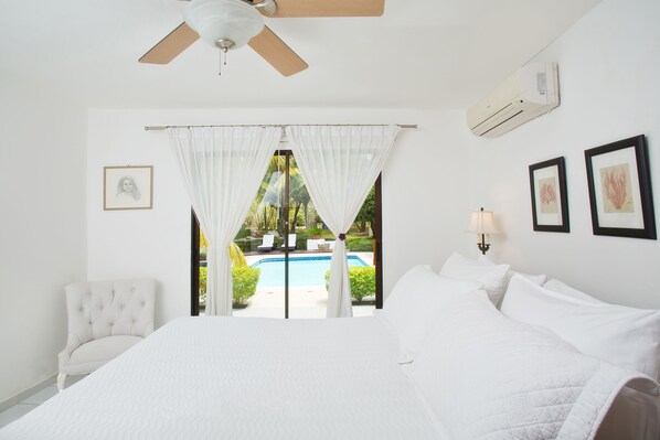 Un dormitorio con una cama grande, ventilador de techo, aire acondicionado, vista a una piscina y una silla.