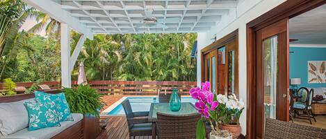 A covered outdoor area with a pool, wooden furniture, and potted plants.