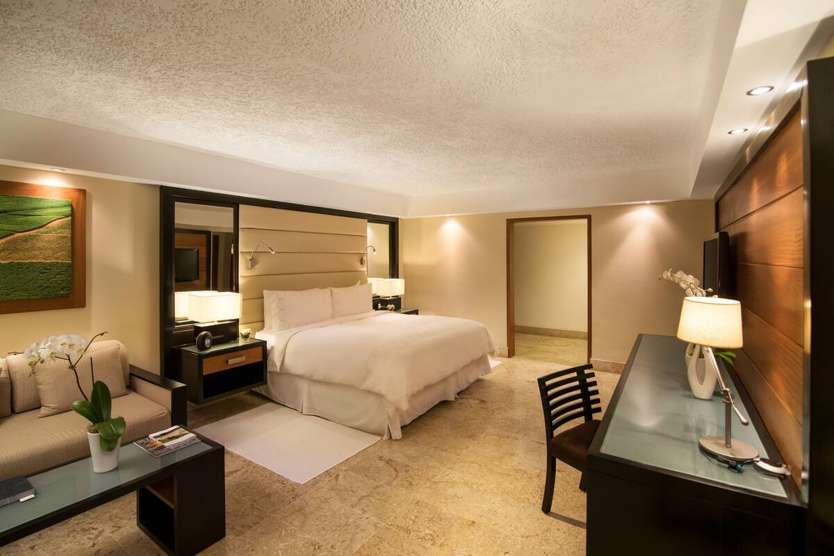 Una habitación de hotel con cama, escritorio, silla, mesita de noche, lámpara, planta y espejo.