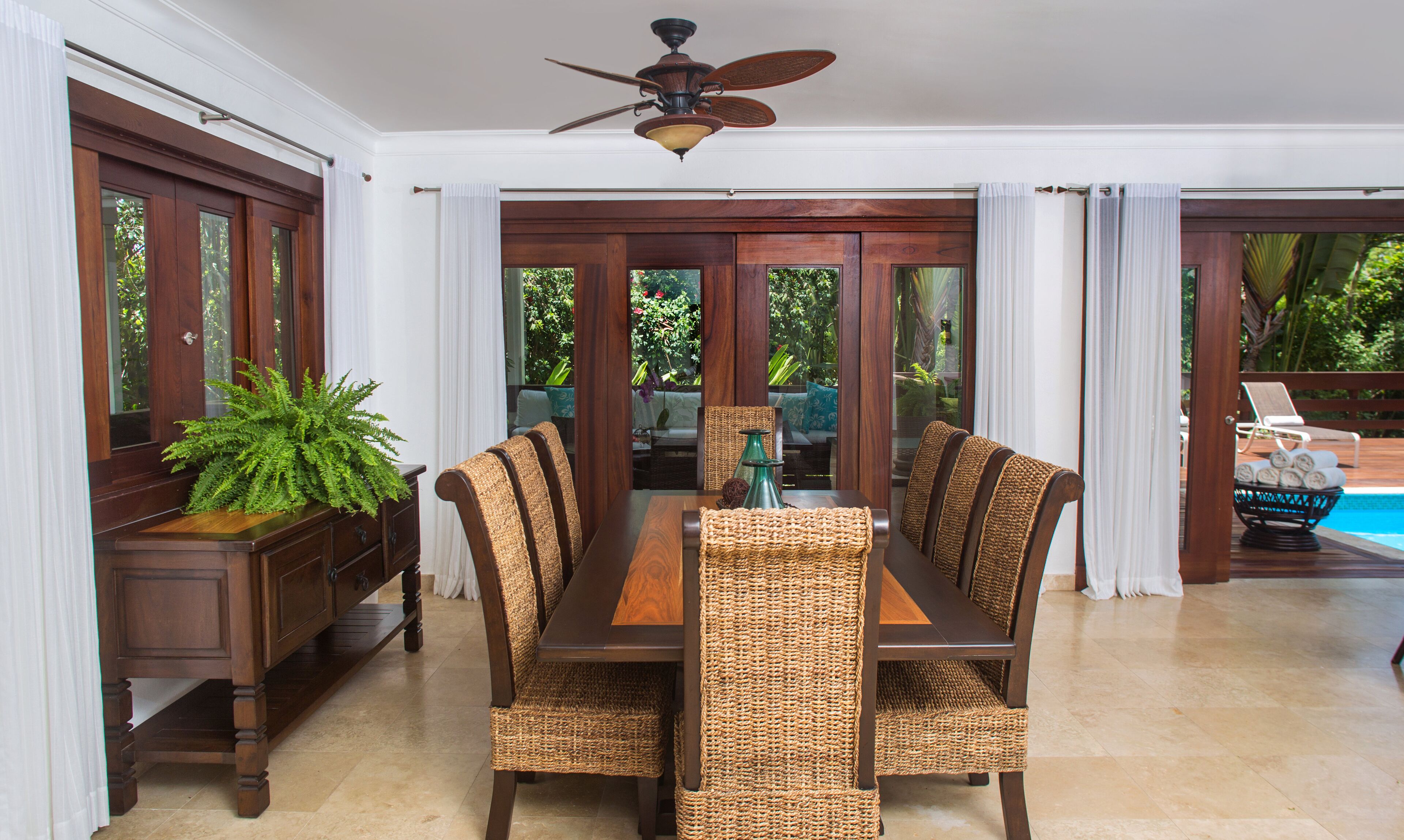 A dining area with a wooden table and wicker chairs, a ceiling fan, and a view of a pool and greenery outside.