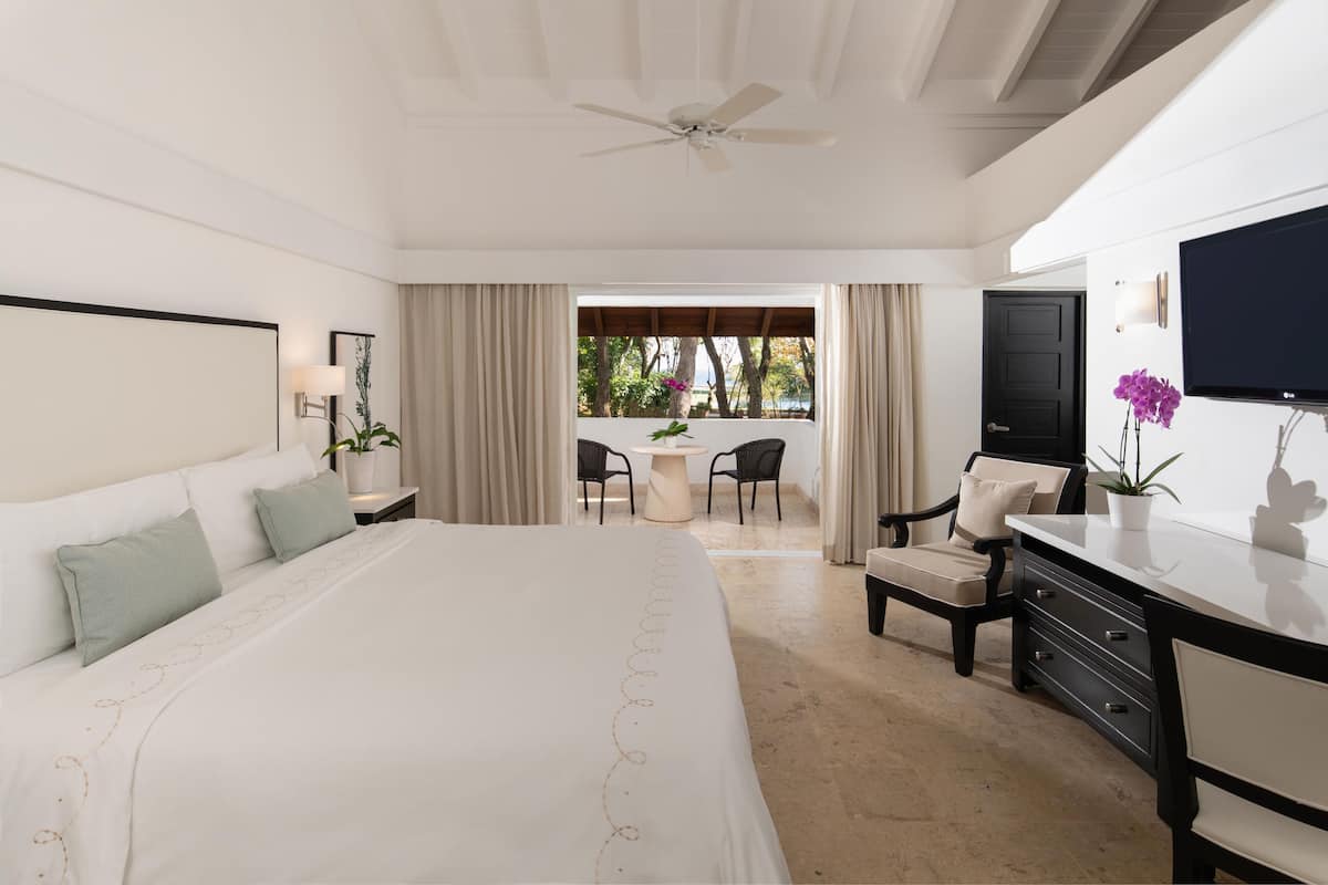 Habitación de hotel con una cama grande, un escritorio, una silla, un televisor y un balcón con mobiliario exterior.