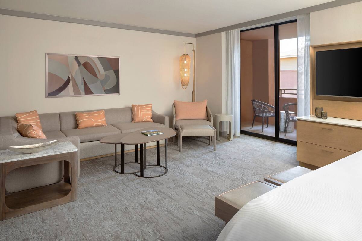 A modern hotel room with a sofa, armchair, small round table, and a bed. There is a television and a window with curtains.