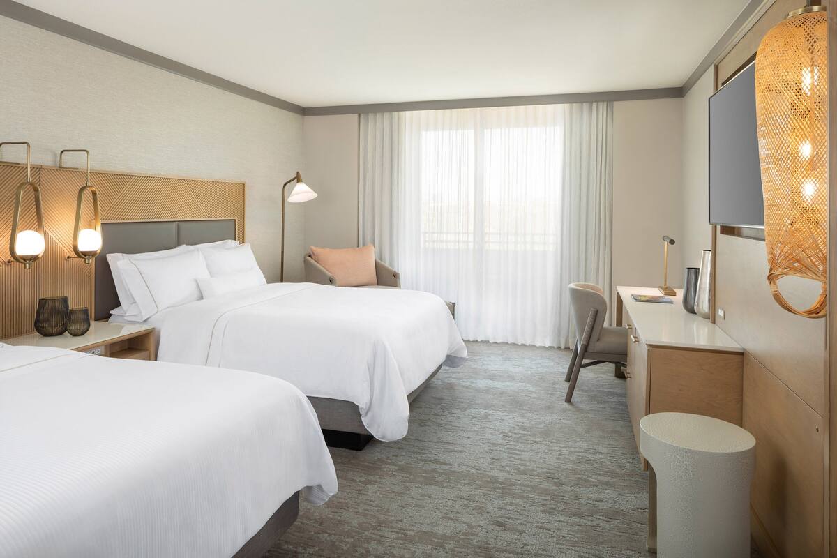 A hotel room with two beds, a desk, a chair, a TV, and a large window with curtains.