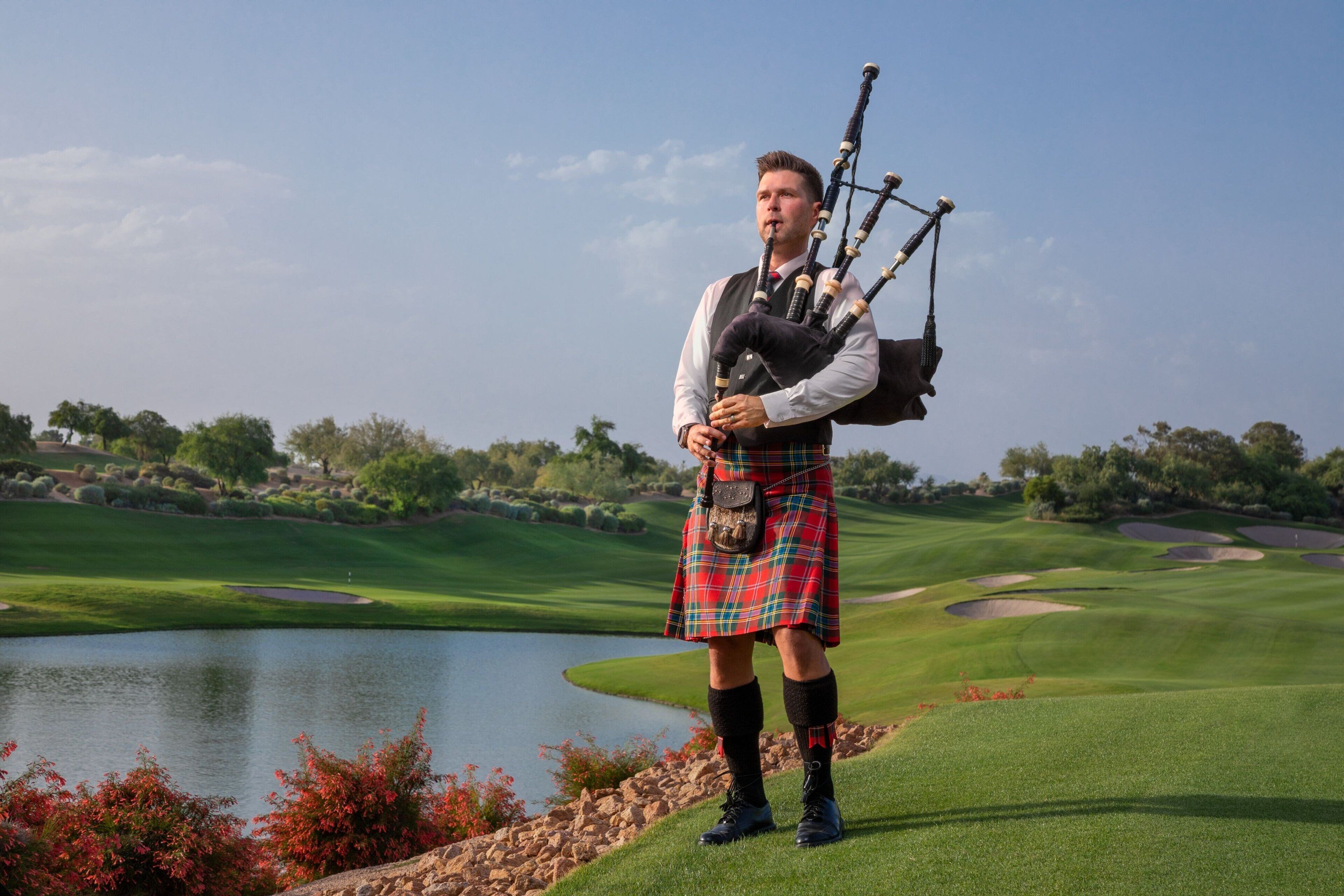 Une personne vêtue d’un costume traditionnel écossais, jouant de la cornemuse sur un terrain de golf, avec un lac et des arbres en arrière-plan.