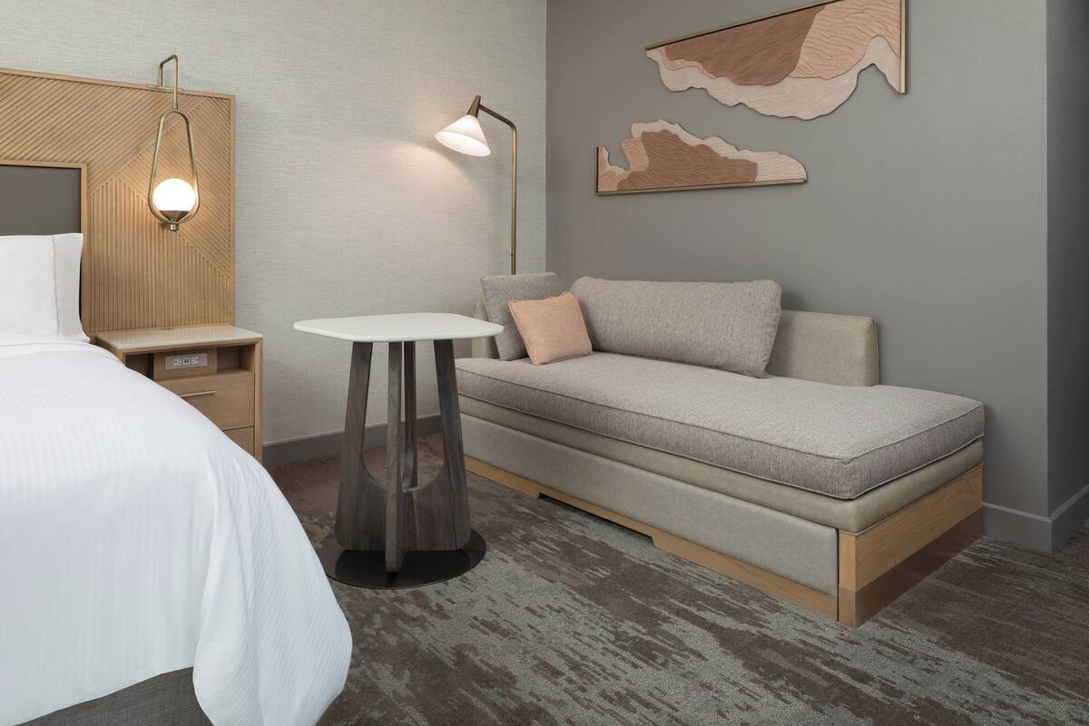 A modern hotel room with a bed, a sofa, a small table, and a lamp.
