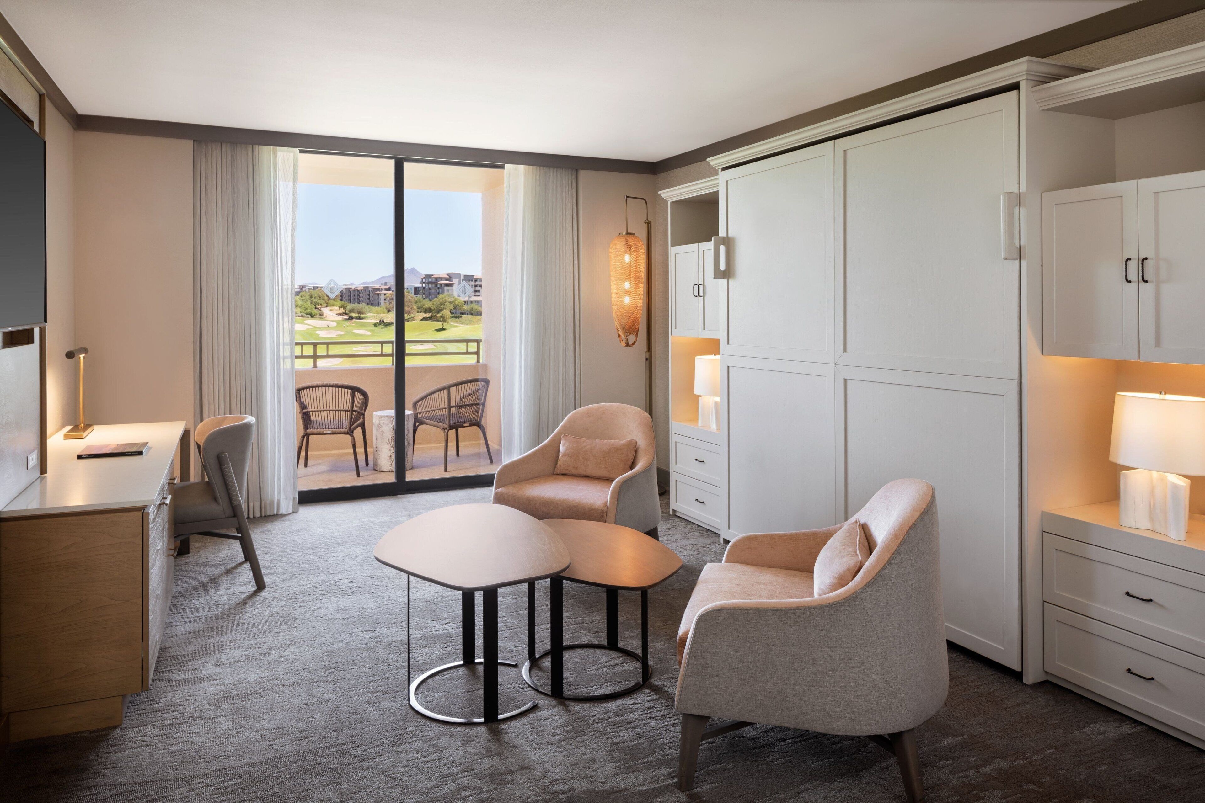Een moderne hotelkamer met een grote schuifpui naar een balkon, een bureau met stoel, twee fauteuils, een kleine tafel en een kledingkast.