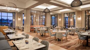 A modern restaurant with multiple dining areas, including a bar and private booths. Tables are set with white tablecloths and silverware.