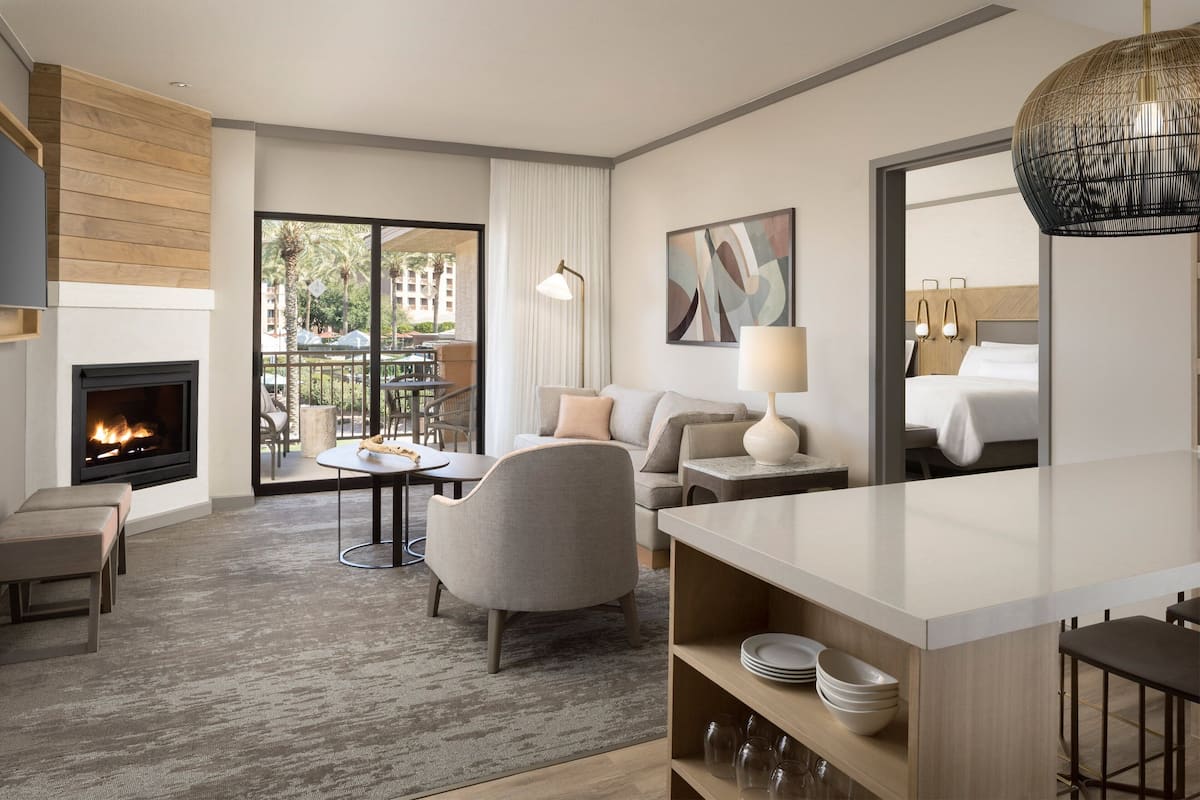 A modern hotel room with a fireplace, a balcony with outdoor furniture, a sofa, a dining table, and a large mirror.
