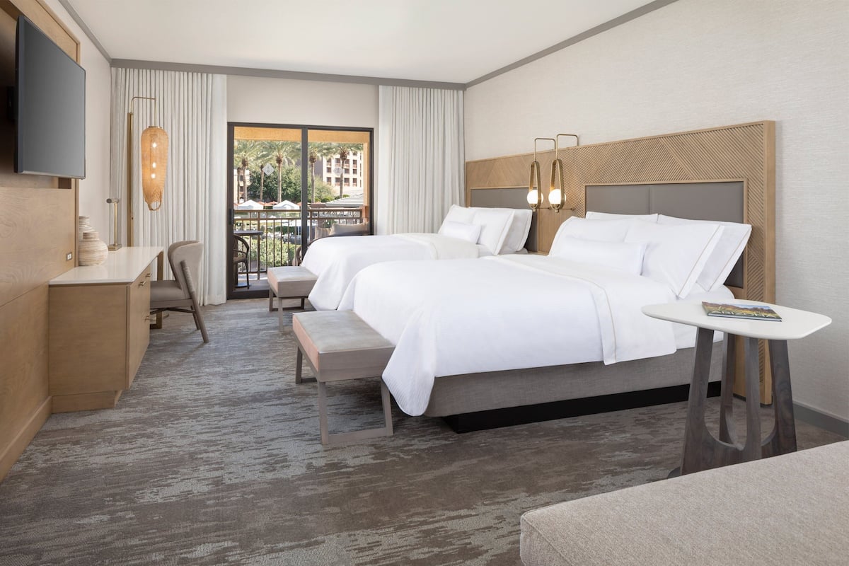 A hotel room with two beds, a desk, a TV, and a balcony with a view of buildings and palm trees.