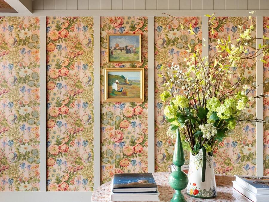A room with floral wallpaper, a framed painting, a vase with flowers, and books on a table.