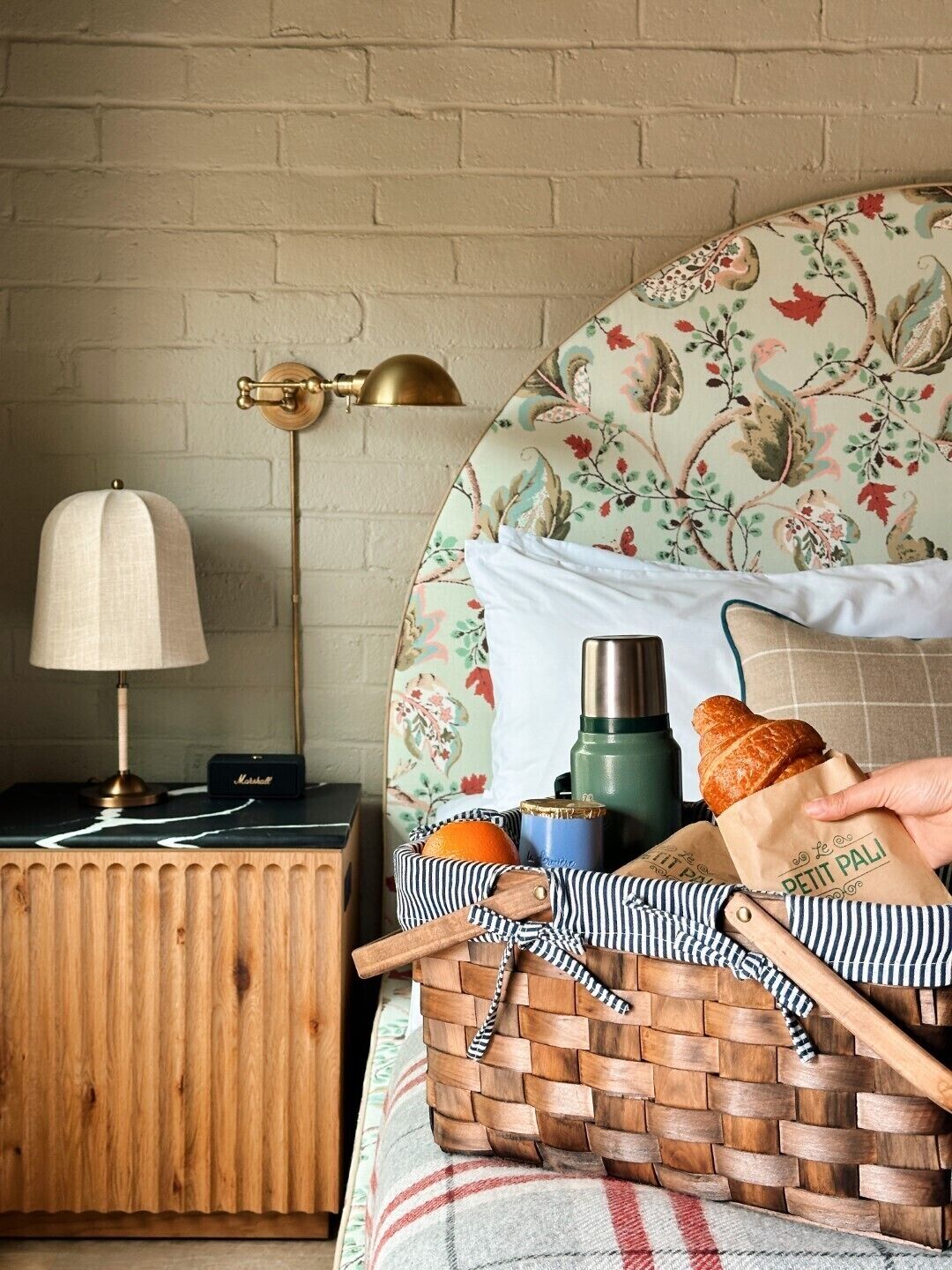 A cozy room with a wooden nightstand, a lamp, a woven basket with a baguette, a bottle, and a paper bag.