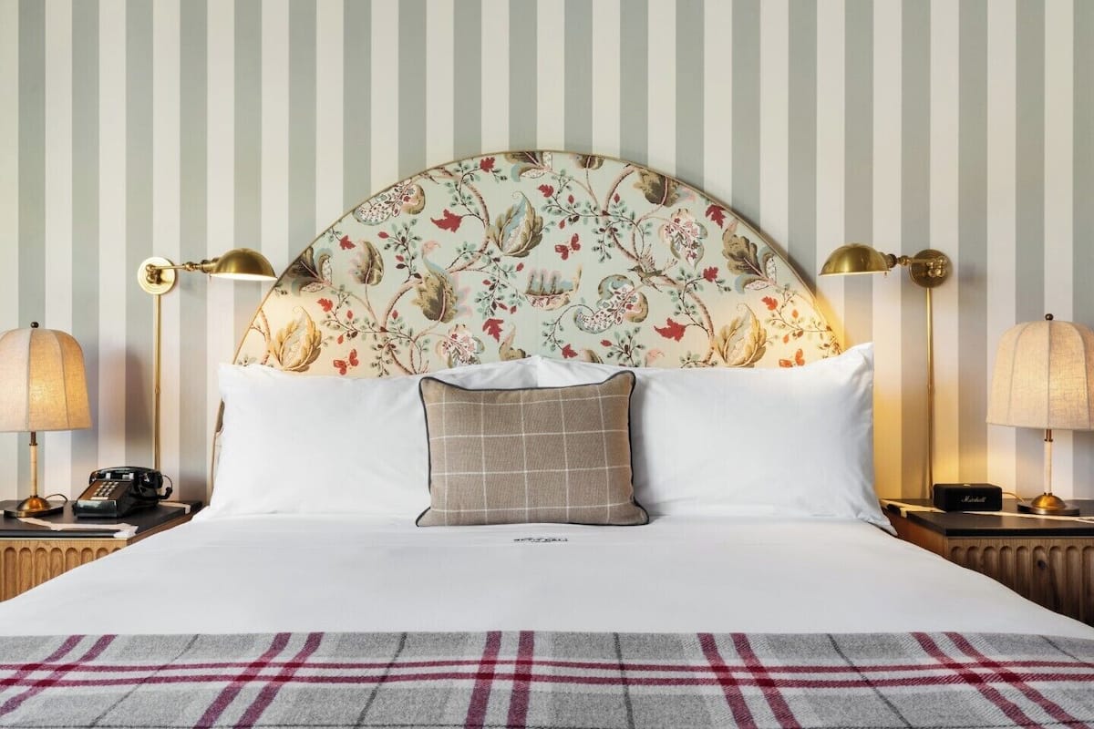 A hotel room with a striped wallpaper, a floral headboard, two bedside lamps, a telephone, and a plaid throw blanket.