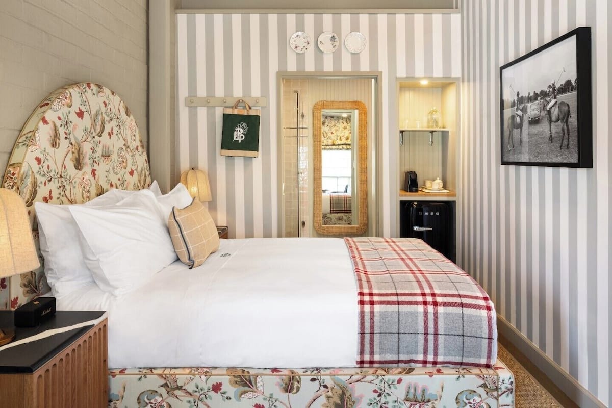 A hotel room with a large bed, a bedside table with a lamp, a dresser, a mirror, and a framed photograph on the wall.