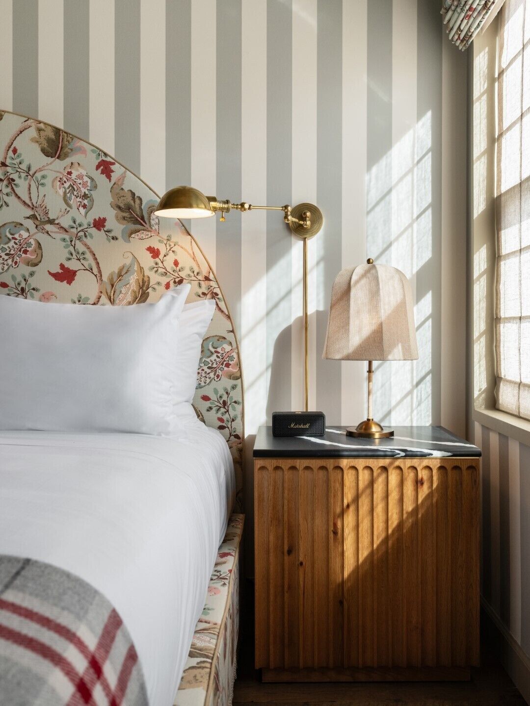 Un dormitorio con cama, mesita de noche, lámpara y ventana con persianas verticales.
