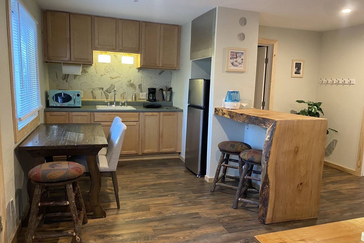 A kitchen with wooden cabinets, a stainless steel refrigerator, a microwave, and a coffee maker. There is a wooden table with two stools and a small wooden island with a sink.