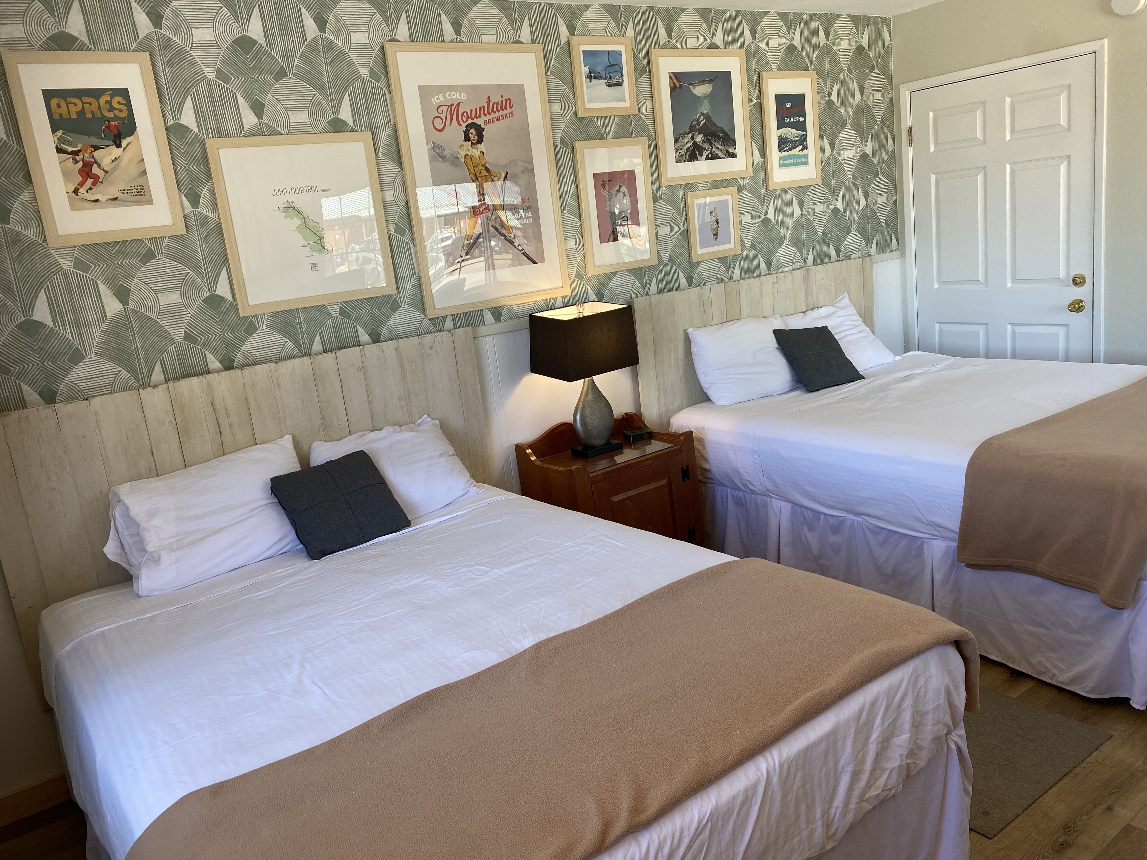 A hotel room with two beds, a wooden headboard, a nightstand with a lamp, and a door.