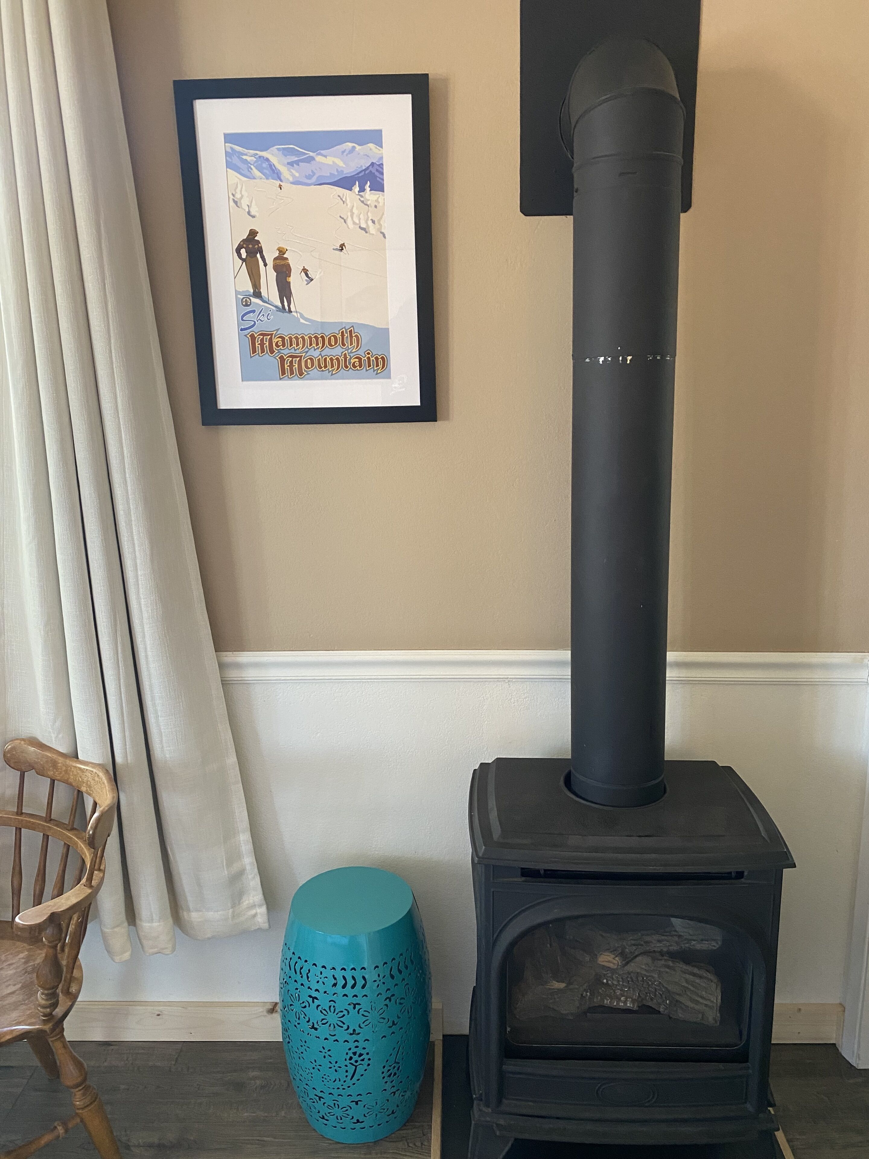 A room with a black wood-burning stove, a framed picture of a mountain scene, a wooden chair, and a teal decorative vase.