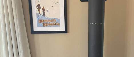 A room with a black wood-burning stove, a framed picture of a mountain scene, a wooden chair, and a teal decorative vase.