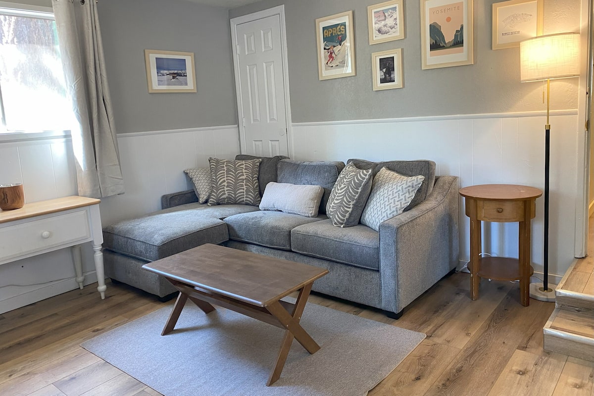 A cozy living room with a grey sectional sofa, a wooden coffee table, and a small round side table. There are framed pictures on the wall, a white door, and a window with curtains.