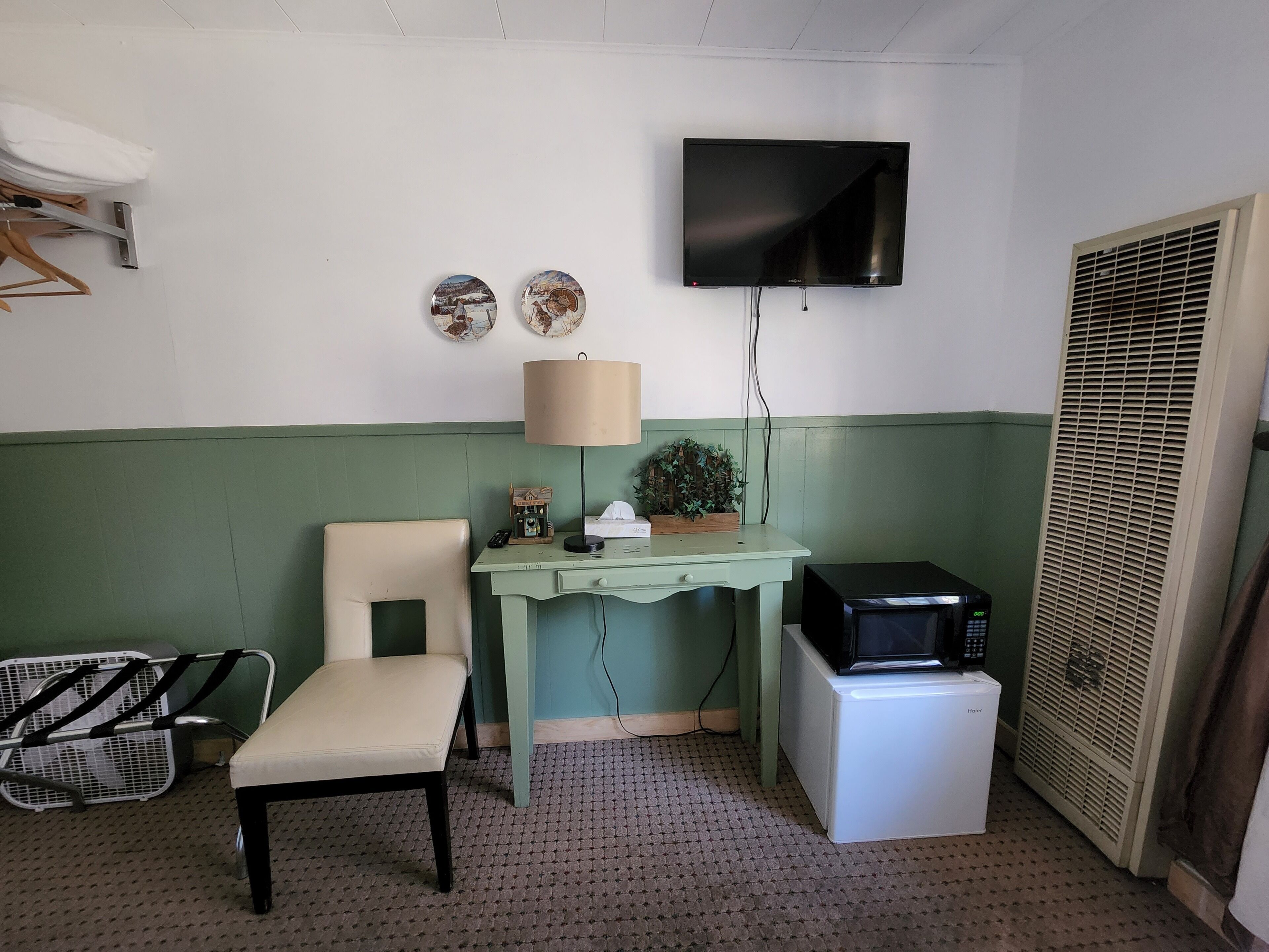 Une petite pièce aux couleurs vert et blanc, avec un téléviseur fixé au mur, un bureau avec une lampe, une chaise, un petit réfrigérateur et un radiateur.
