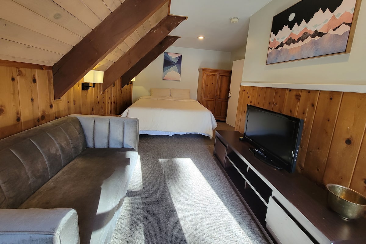 A cozy room with a wooden ceiling, a sofa, a bed, a TV, and a painting on the wall.