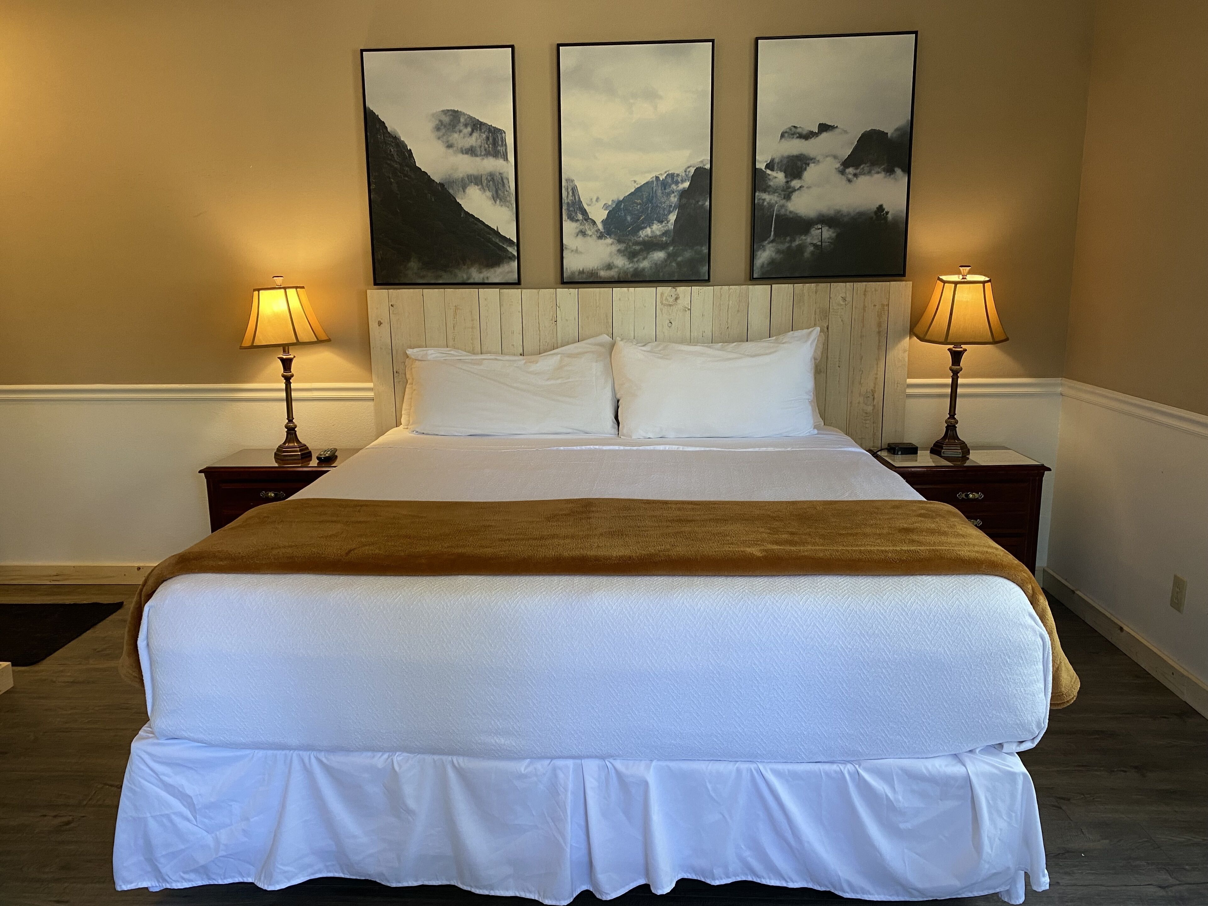 A neatly made bed with white linens and a brown throw, flanked by two bedside tables with lamps, against a wall with three framed mountain landscape pictures.