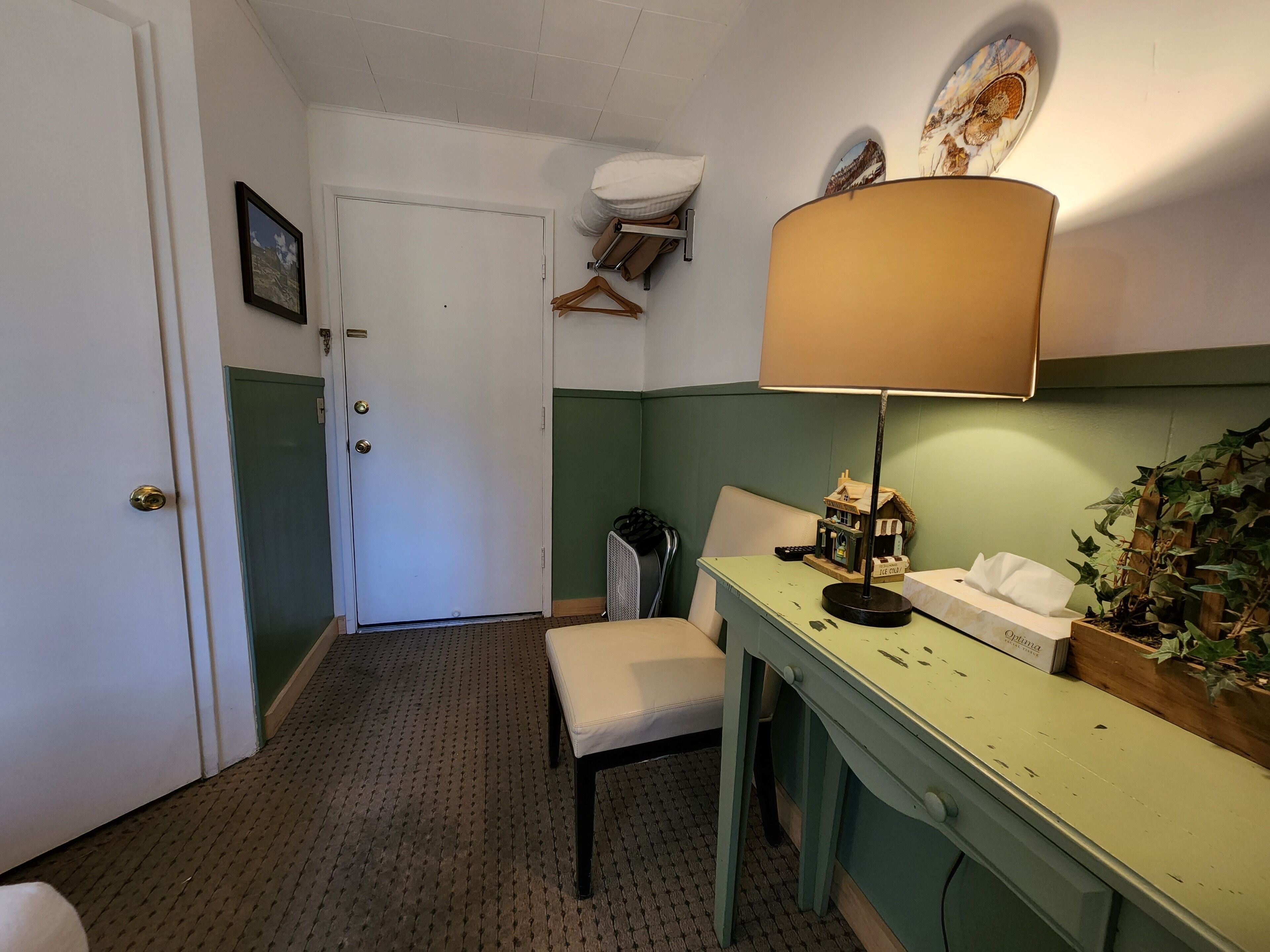 Une petite pièce accueillante aux couleurs vertes et blanches, avec un seul fauteuil, un bureau doté d’une lampe, une petite plante et une étagère fixée au mur, sur laquelle reposent une télécommande et quelques objets décoratifs.