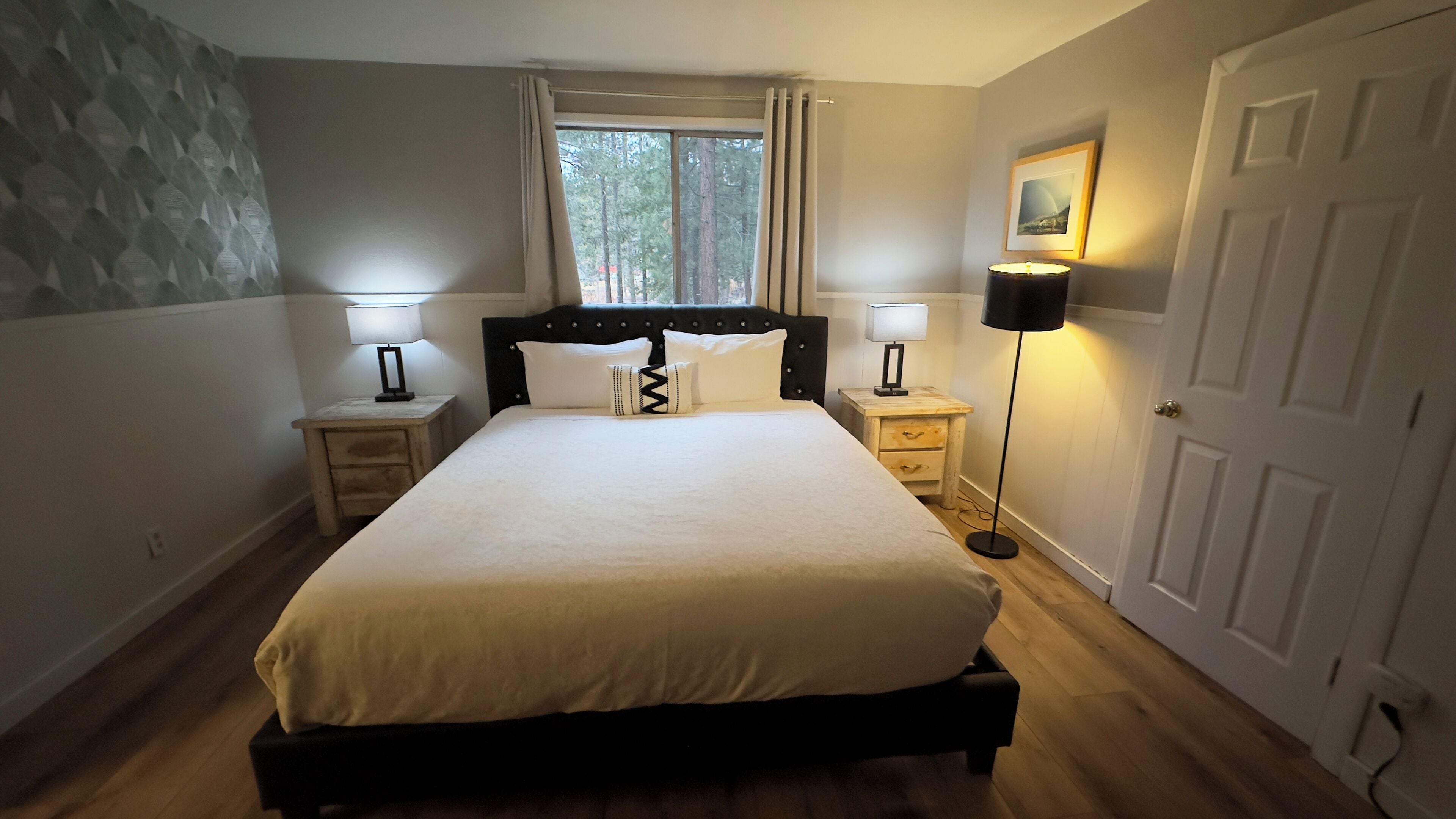 A bedroom with a large bed, two bedside tables with lamps, a window with curtains, a door, and a wall with a textured pattern.