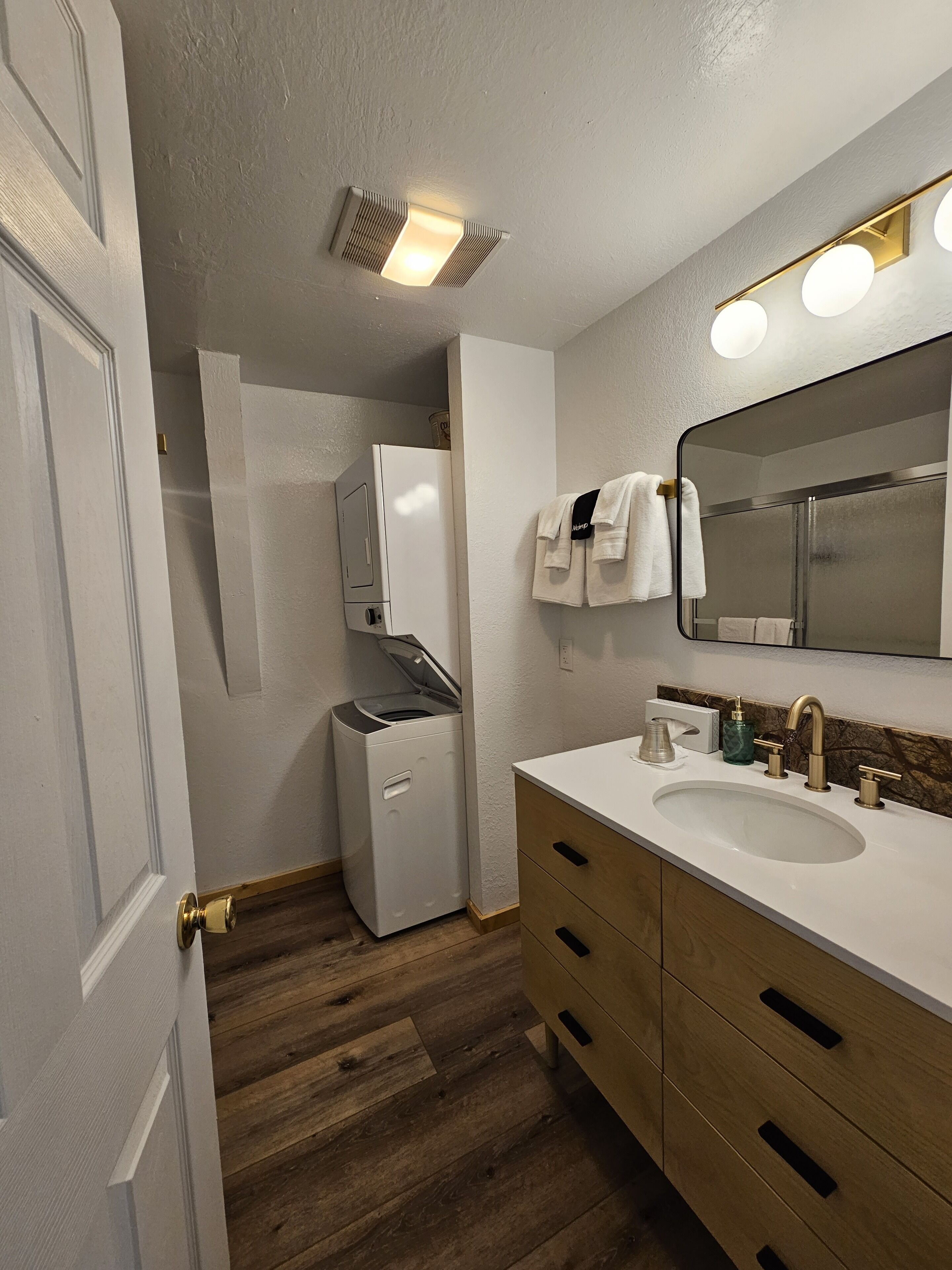A bathroom with a white sink, wooden vanity, a mirror with lights, a towel rack, and a washing machine.