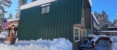 A green building with a small window and a door, surrounded by snow and trees.