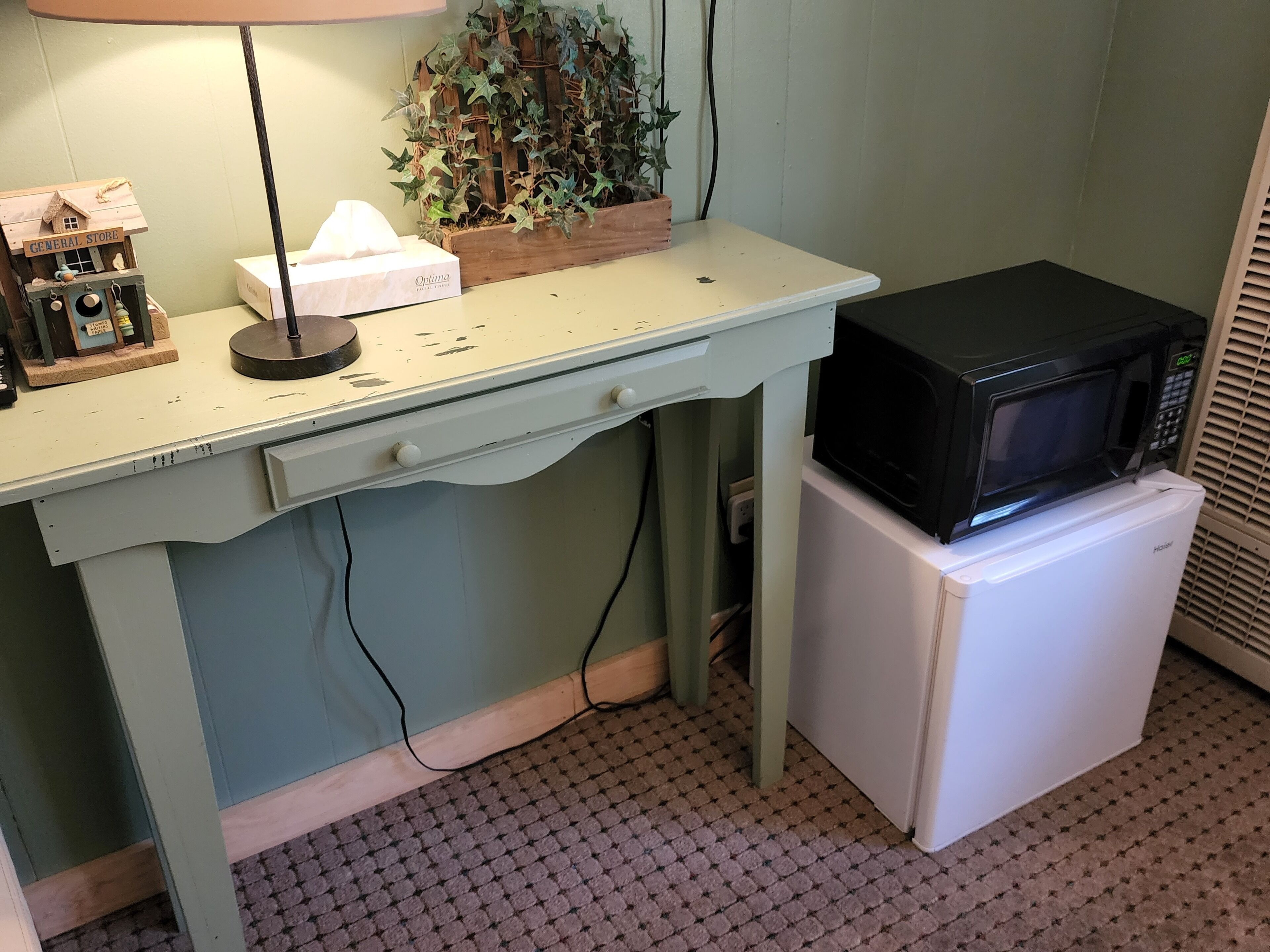 A small kitchenette with a white refrigerator, a microwave, a wooden shelf with a plant, a lamp, and a small toy house.