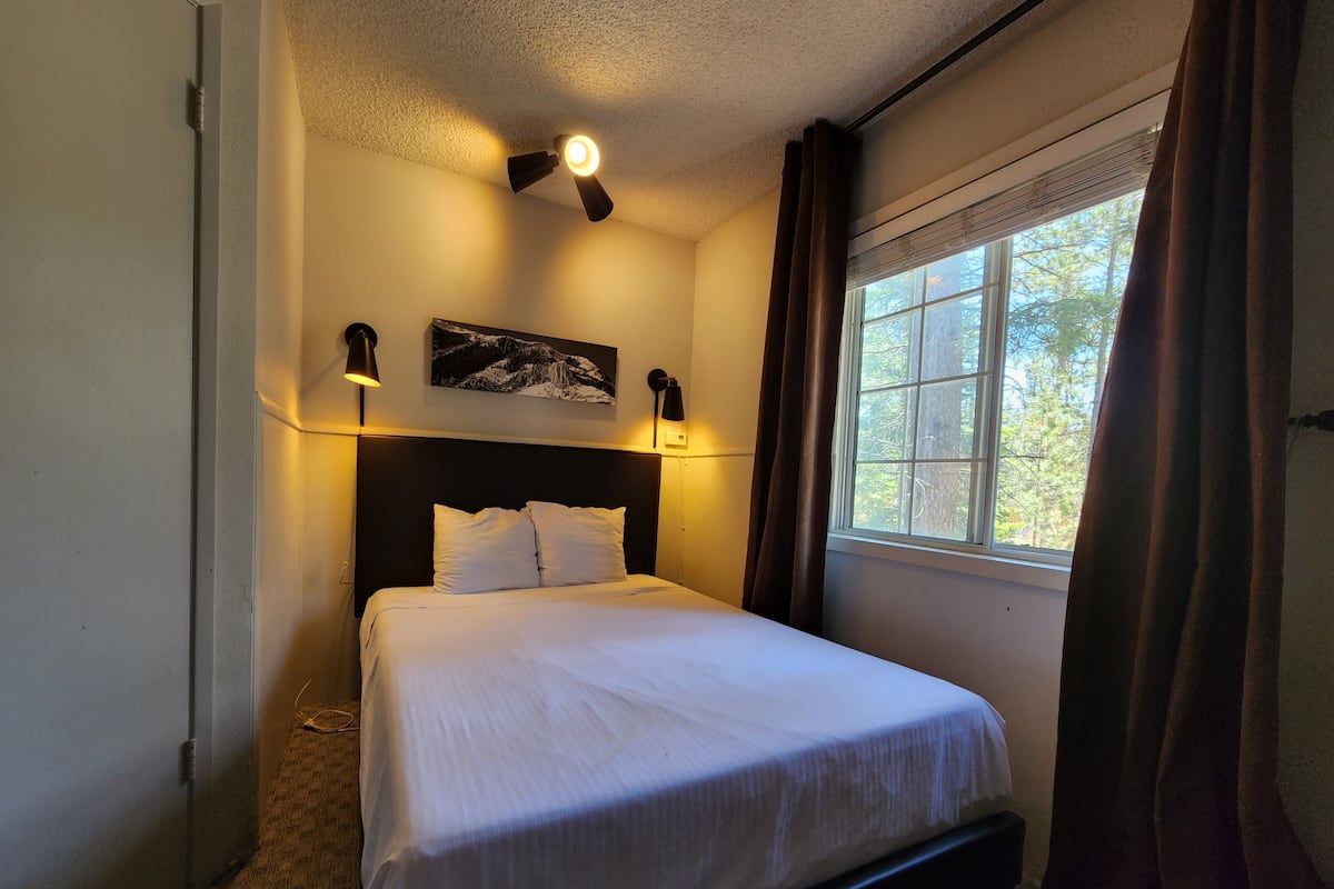 A neatly made bed with white linens, a dark headboard, and two wall-mounted lamps. A window with curtains allows natural light and a view of trees outside.